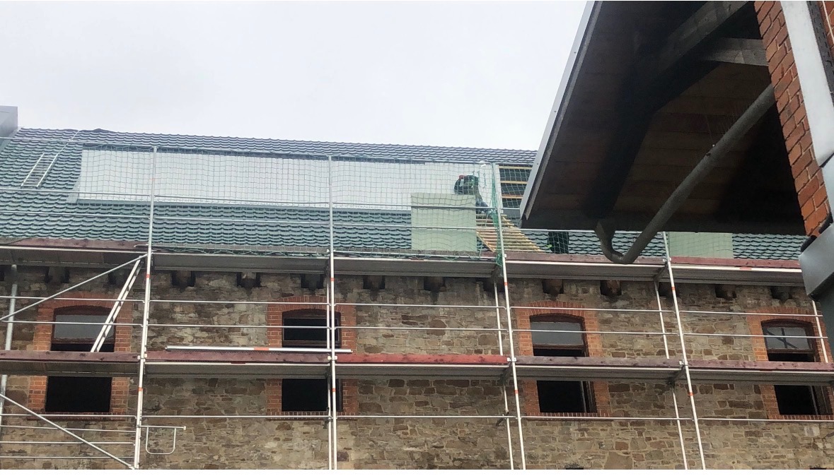 Budynek w rusztowaniach z zabezpieczeniem na dachu podczas prac. Widoczny fragment rynny i okna. Dachówka w odcieniach szarości i zieleni.