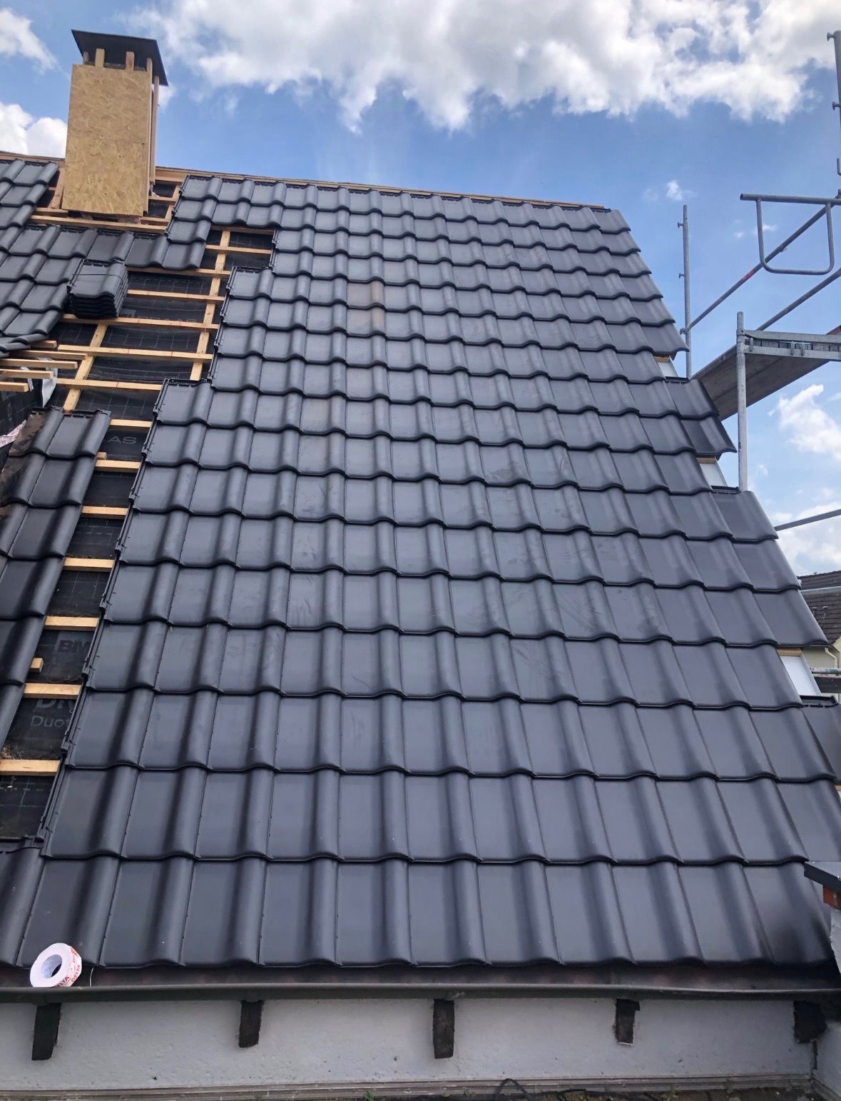 Częściowo pokryty dach czarną dachówką ceramiczną, widoczna więźba i komin w trakcie budowy, rusztowanie z boku. Niebieskie niebo z chmurami w tle.