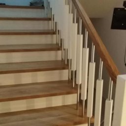 Almar - Drewniane schody z białymi balustradami i metalowymi elementami, widok z dołu. Naturalne oświetlenie podkreśla strukturę drewna i nowoczesny design.