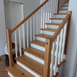 Almar - Montaż drewnianych schodów z białymi balustradami w jasnym wnętrzu. Widoczny stolarz podczas pracy, narzędzia i elementy wykończeniowe. Naturalne oświetlenie.