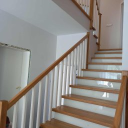 Almar - Drewniane schody z białymi balustradami i podstopnicami z mlecznego szkła, widok z dołu. Naturalne oświetlenie, minimalistyczny design. Detal wykończenia wnętrza.