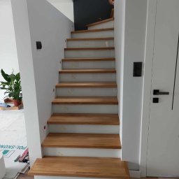 Almar - Nowoczesne schody drewniane z białymi elementami konstrukcyjnymi, widok z dołu. Naturalne oświetlenie, minimalistyczny design, widoczny fragment osoby na górze schodów.