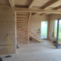 Almar - Drewniane schody w surowym wnętrzu domu z boazerią, widok na otwarte okno i drzwi. Żółta miotła oparta o ścianę. Wnętrze w trakcie wykańczania.