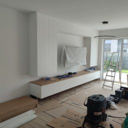 Almar - Biała zabudowa meblowa w salonie, z drewnianym blatem, częściowo zakryta folią ochronną. Widoczne narzędzia, drabina i odkurzacz przemysłowy. Podłoga zabezpieczona kartonem.