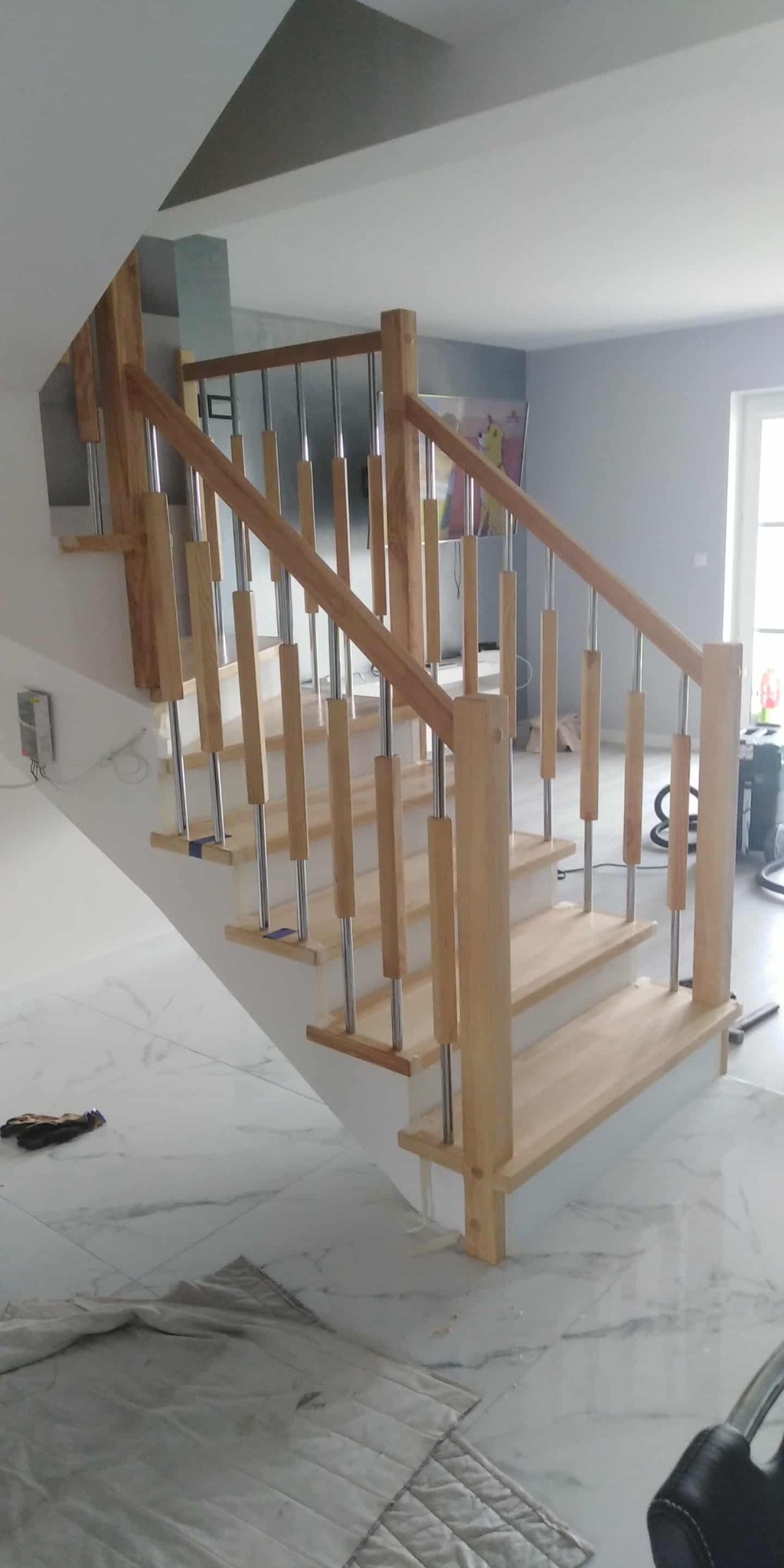 Jasne drewniane schody z metalowymi elementami w nowoczesnym wnętrzu. Widoczne zabezpieczenia na stopniach i narzędzia, sugerujące świeży montaż.