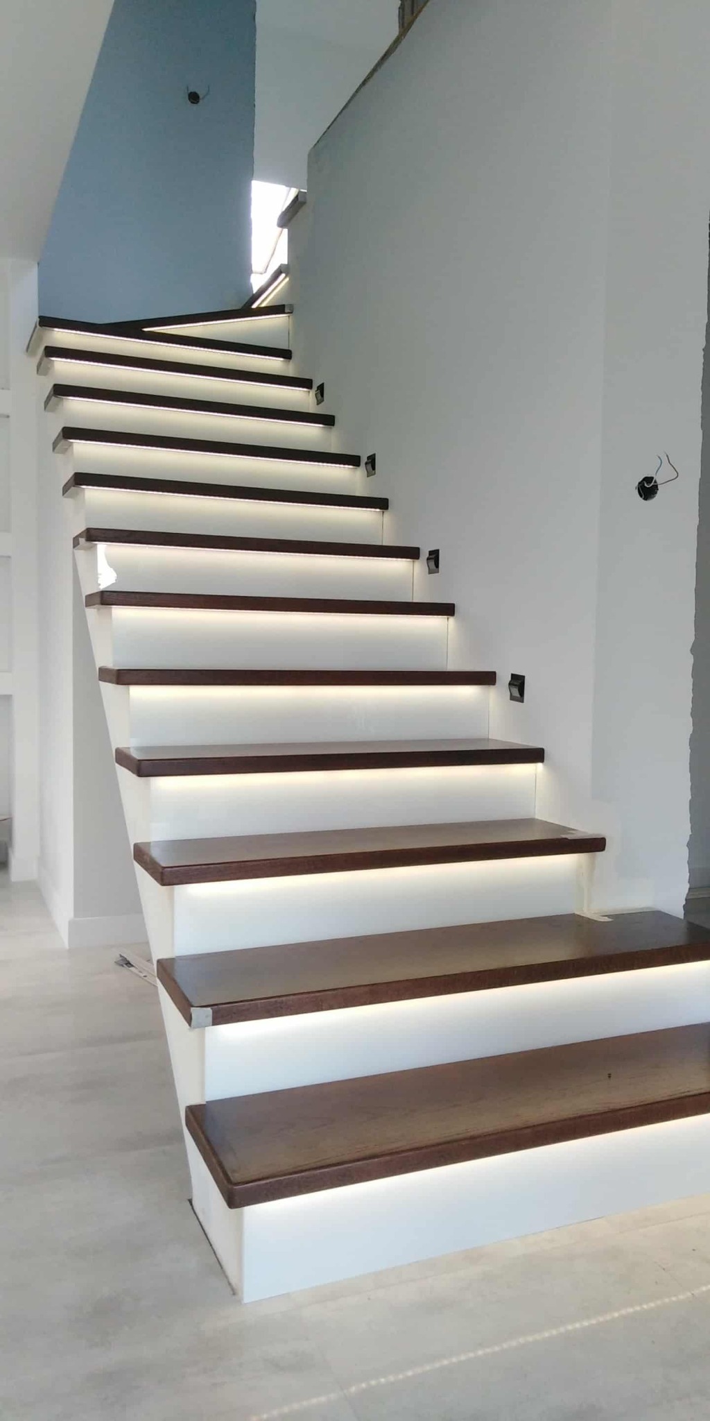 Nowoczesne schody drewniane z podświetleniem LED, widok z dołu. Stopnie w kolorze ciemnego drewna, białe podstopnice. Minimalistyczny design, surowe ściany w tle.