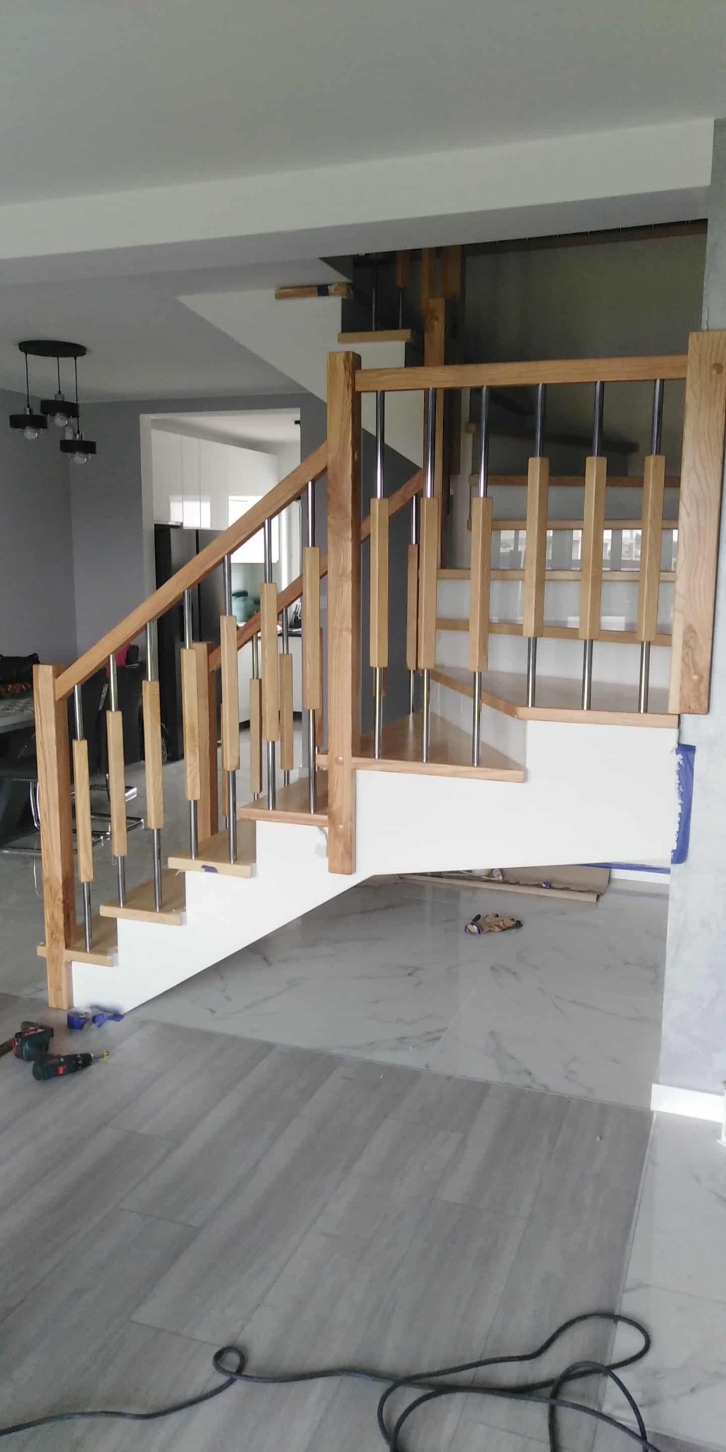 Jasne drewniane schody z metalowymi balustradami w nowoczesnym wnętrzu. Widoczne narzędzia sugerują trwający montaż. Marmurowe płytki podłogowe.