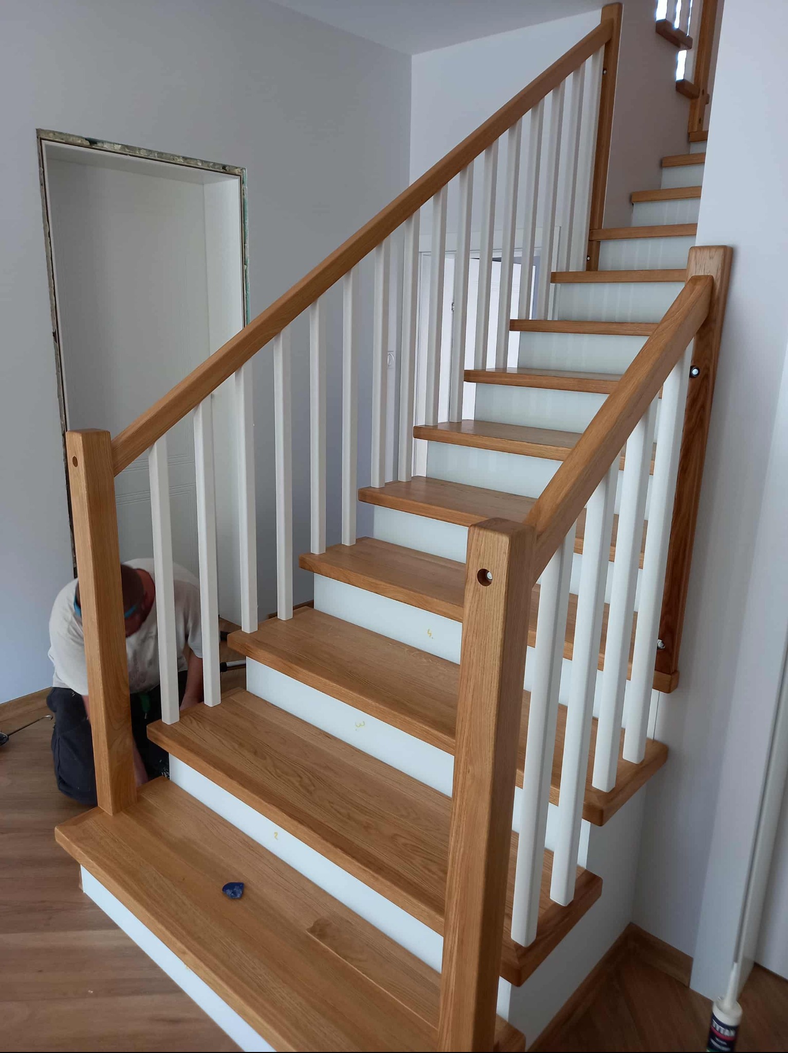 Montaż drewnianych schodów z białymi balustradami w jasnym wnętrzu. Widoczny stolarz podczas pracy, narzędzia i elementy wykończeniowe. Naturalne oświetlenie.