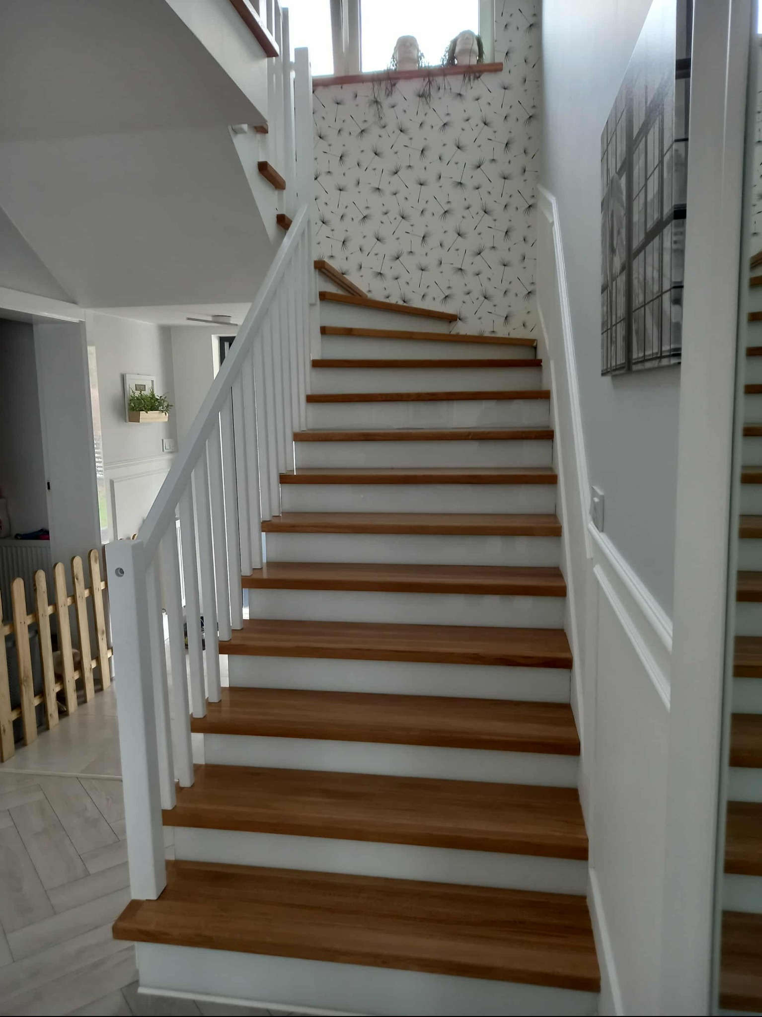 Bieg drewnianych schodów z białymi balustradami w jasnym wnętrzu. Widoczny fragment dekoracyjnej tapety i drewniany płotek zabezpieczający.