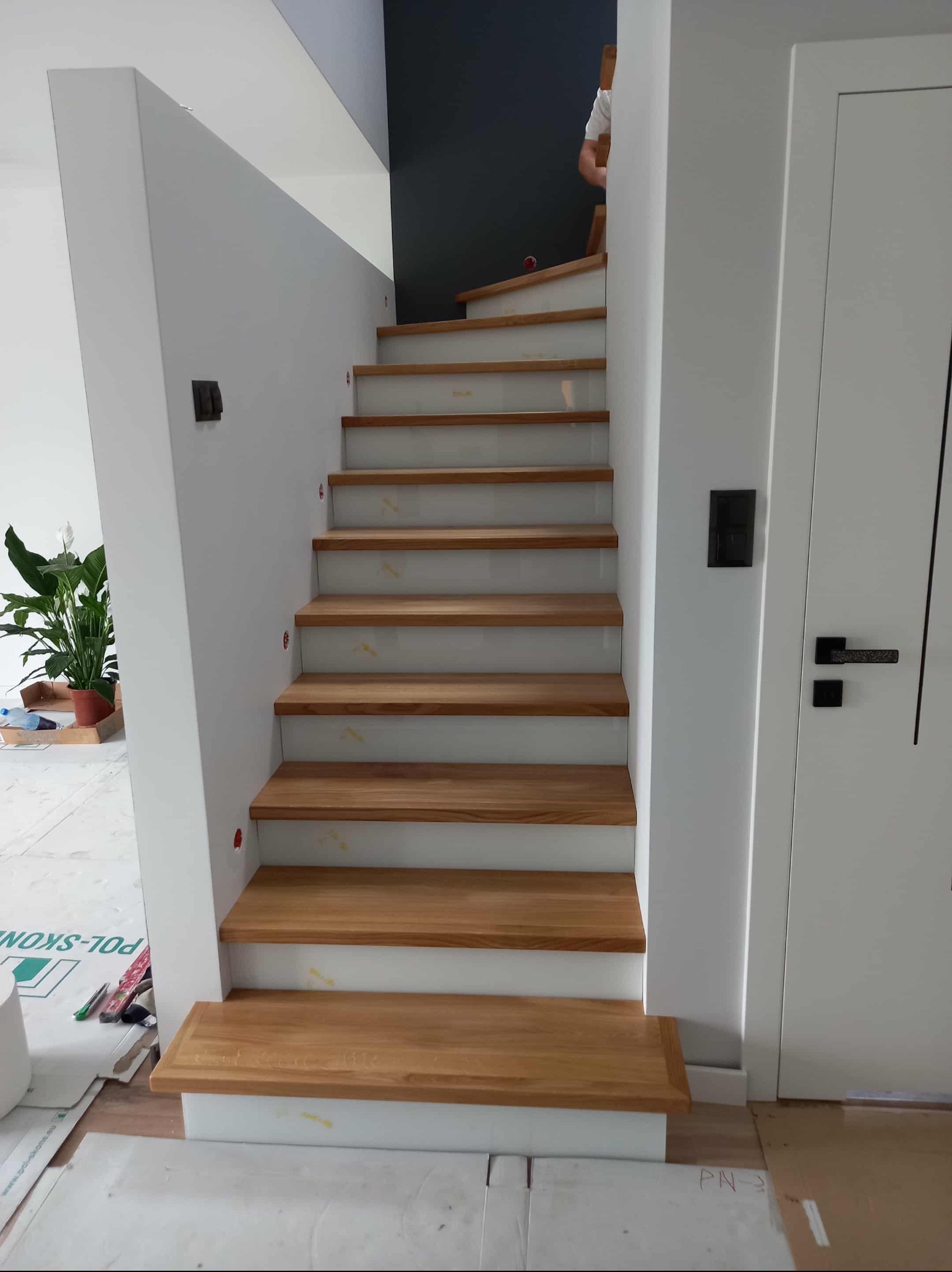 Nowoczesne schody drewniane z białymi elementami konstrukcyjnymi, widok z dołu. Naturalne oświetlenie, minimalistyczny design, widoczny fragment osoby na górze schodów.