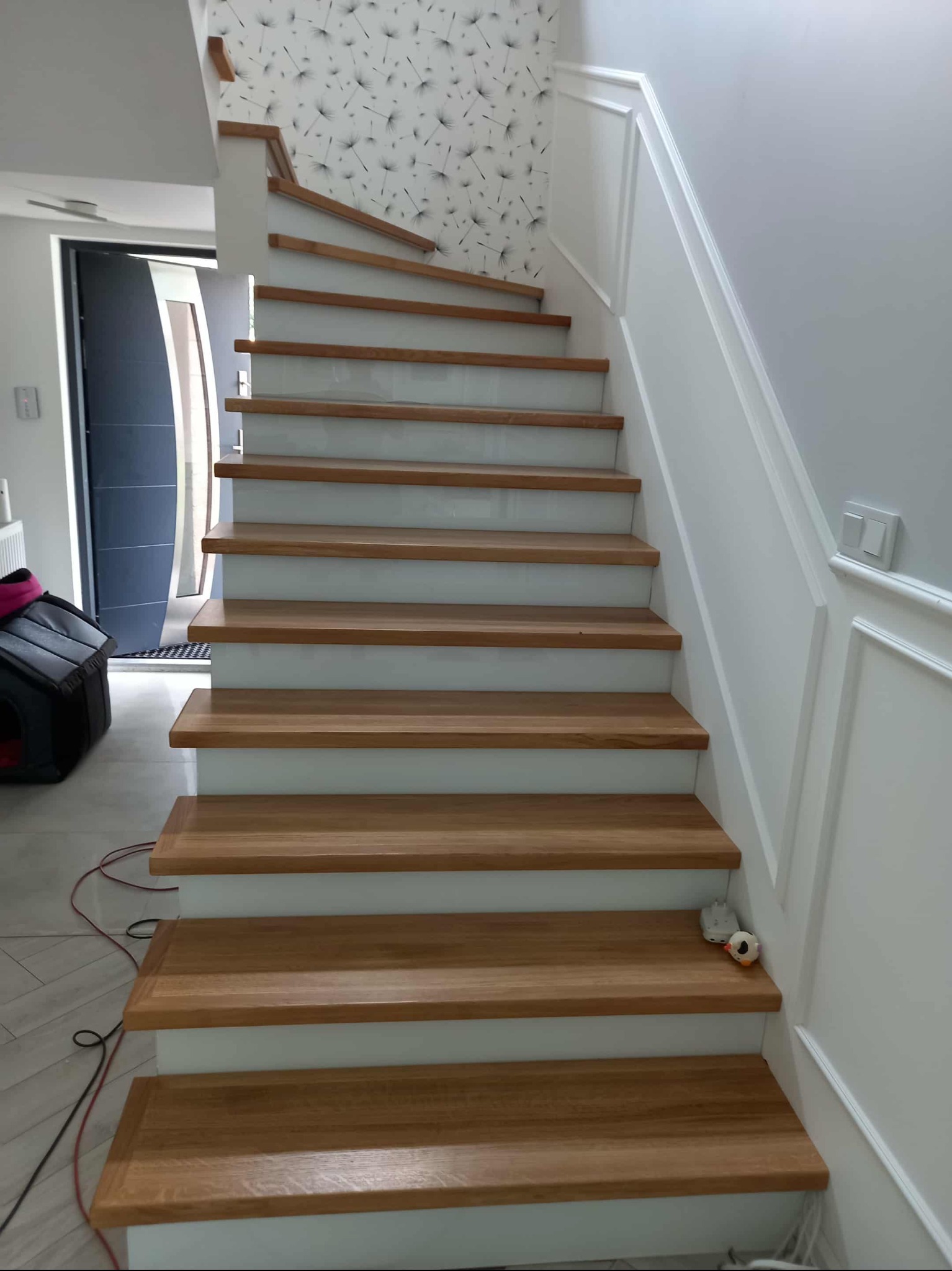 Drewniane schody z białymi podstopnicami i ozdobną listwą ścienną. Widoczny fragment tapety w kwiatowy wzór i czarny transporter dla zwierząt w tle.