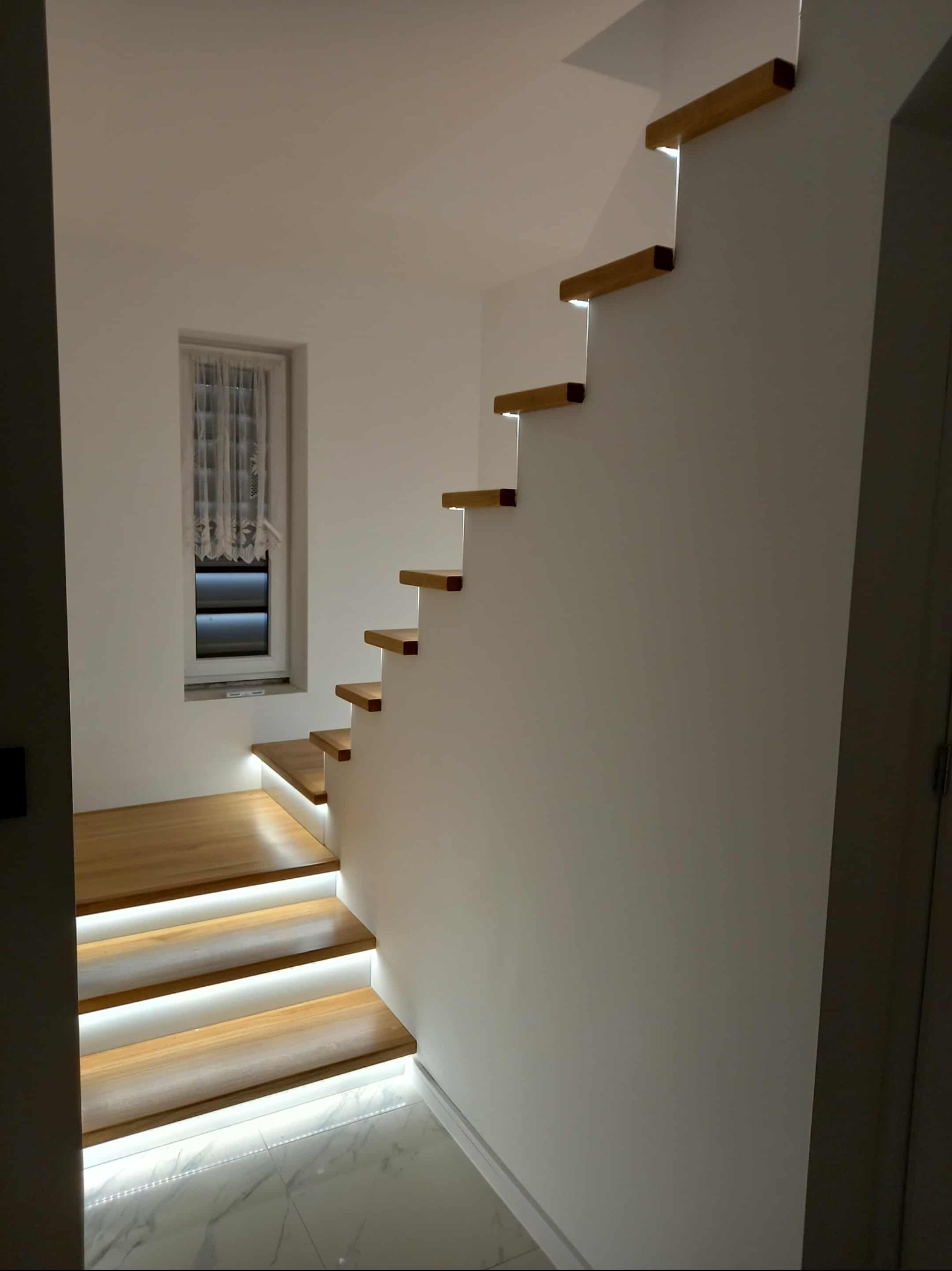 Nowoczesne schody drewniane z podświetleniem LED, zamontowane przy białej ścianie z oknem. Widoczny fragment marmurowej podłogi i drzwi.