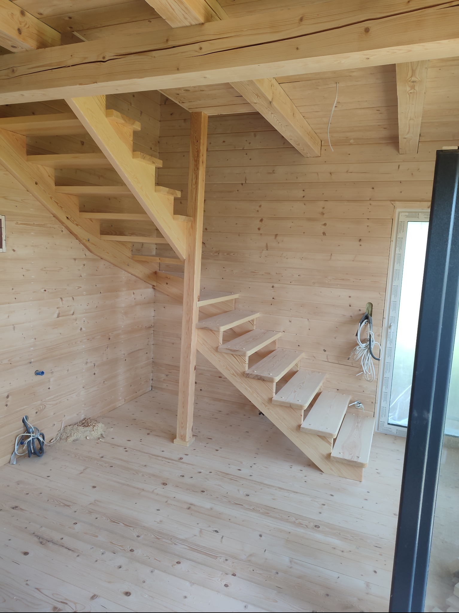 Jasne, drewniane schody spiralne w surowym wnętrzu z boazerią. Widoczne narzędzia i kable, co sugeruje etap montażu. Naturalne światło podkreśla fakturę drewna.