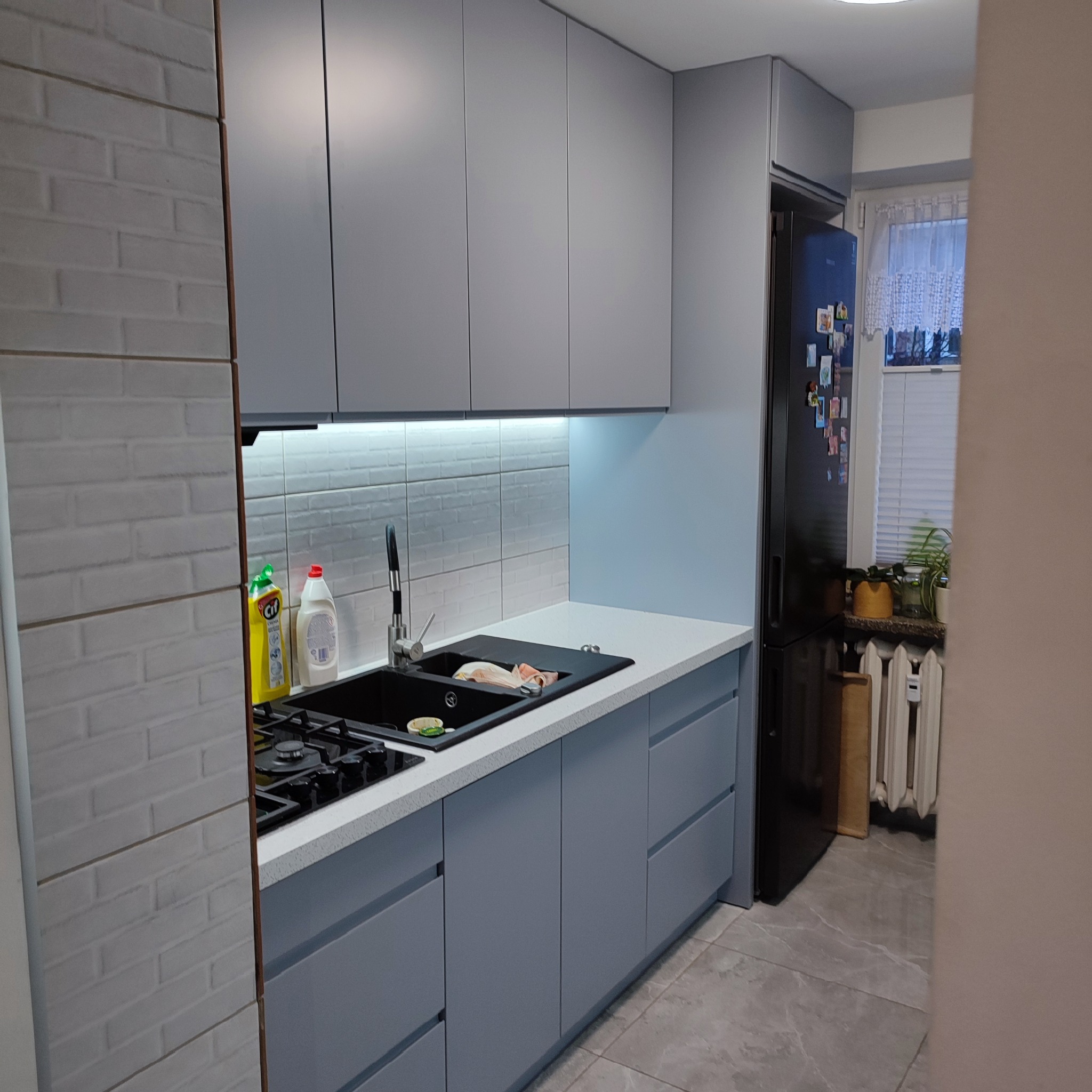 Szara kuchnia na wymiar z czarnym zlewem i płytą. Białe płytki na ścianie i czarna lodówka. Widok na okno z roślinami. Nowoczesny design.