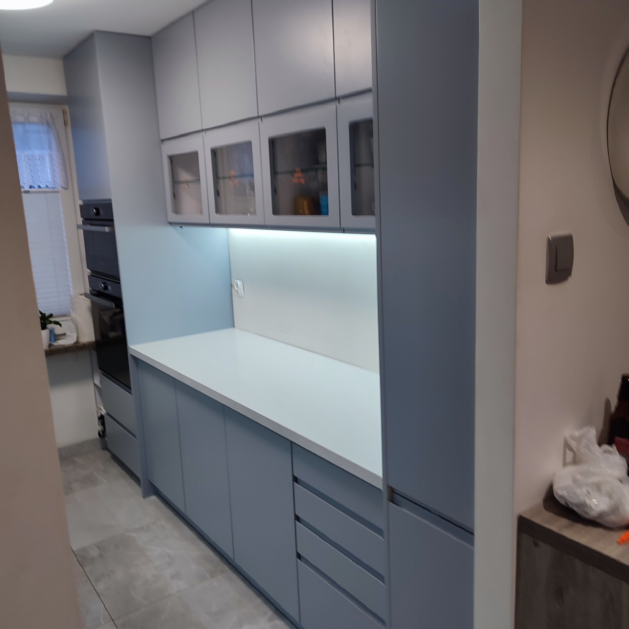 Niebieska kuchnia z białym blatem i przeszklonymi szafkami wiszącymi, oświetlona od spodu. Widok na zabudowę od strony wejścia do pomieszczenia.