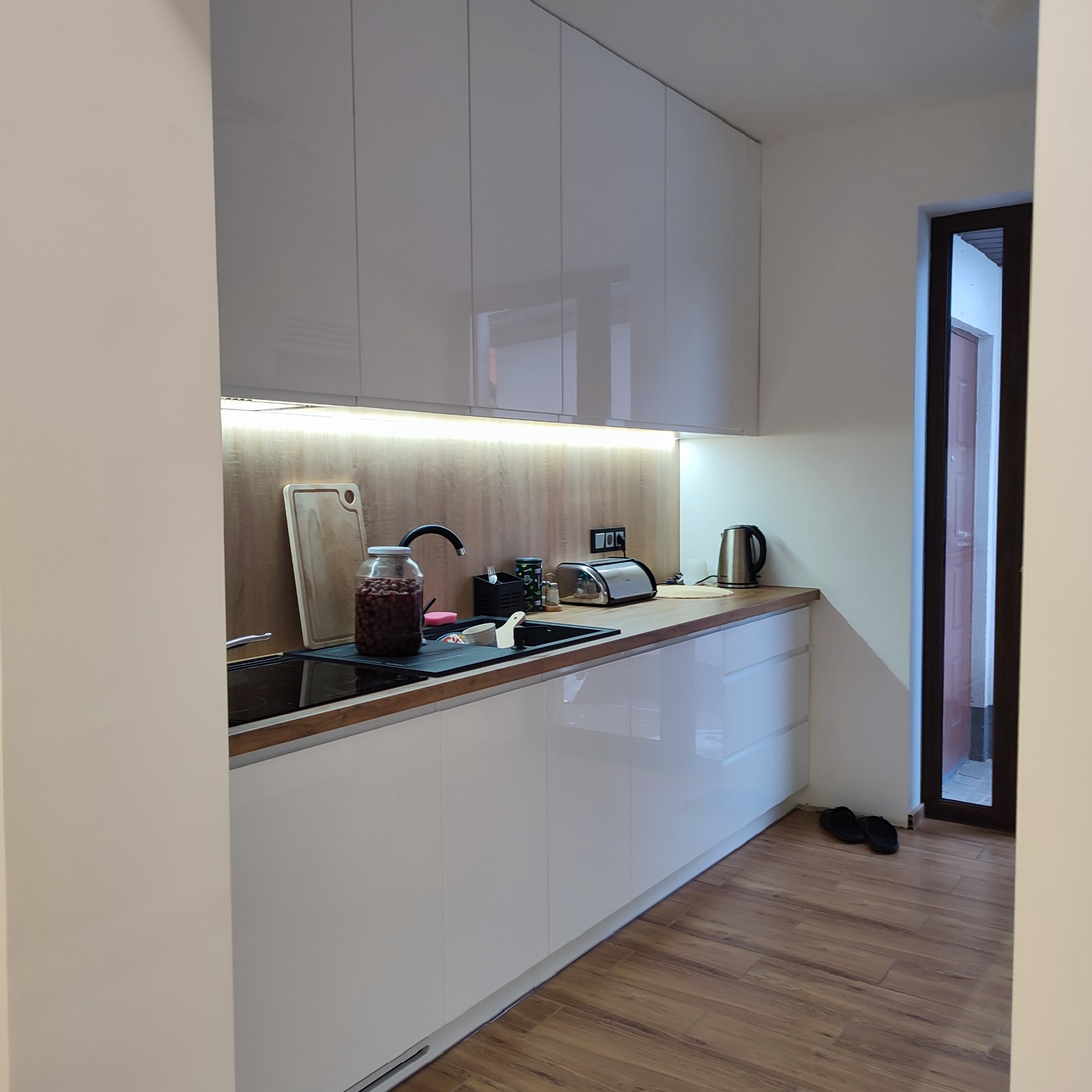 Biała, nowoczesna kuchnia z połyskiem, oświetleniem LED podszafkowym i drewnianym blatem. Widoczny zlew, płyta indukcyjna, czajnik i akcesoria kuchenne. Podłoga drewniana.
