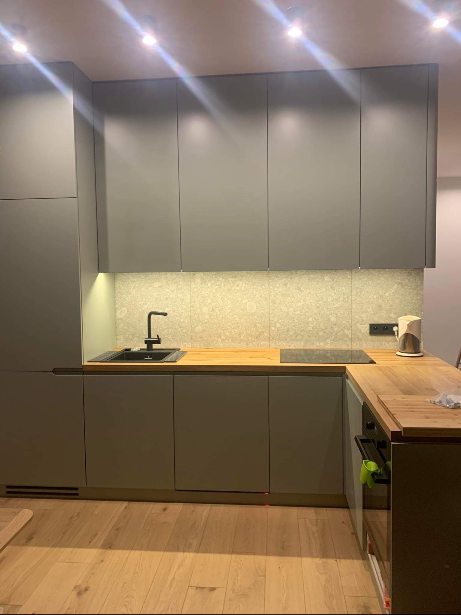 Szara kuchnia z drewnianym blatem i płytkami imitującymi kamień. Nowoczesne oświetlenie podszafkowe. Minimalistyczny design z czarnym zlewem i płytą indukcyjną.