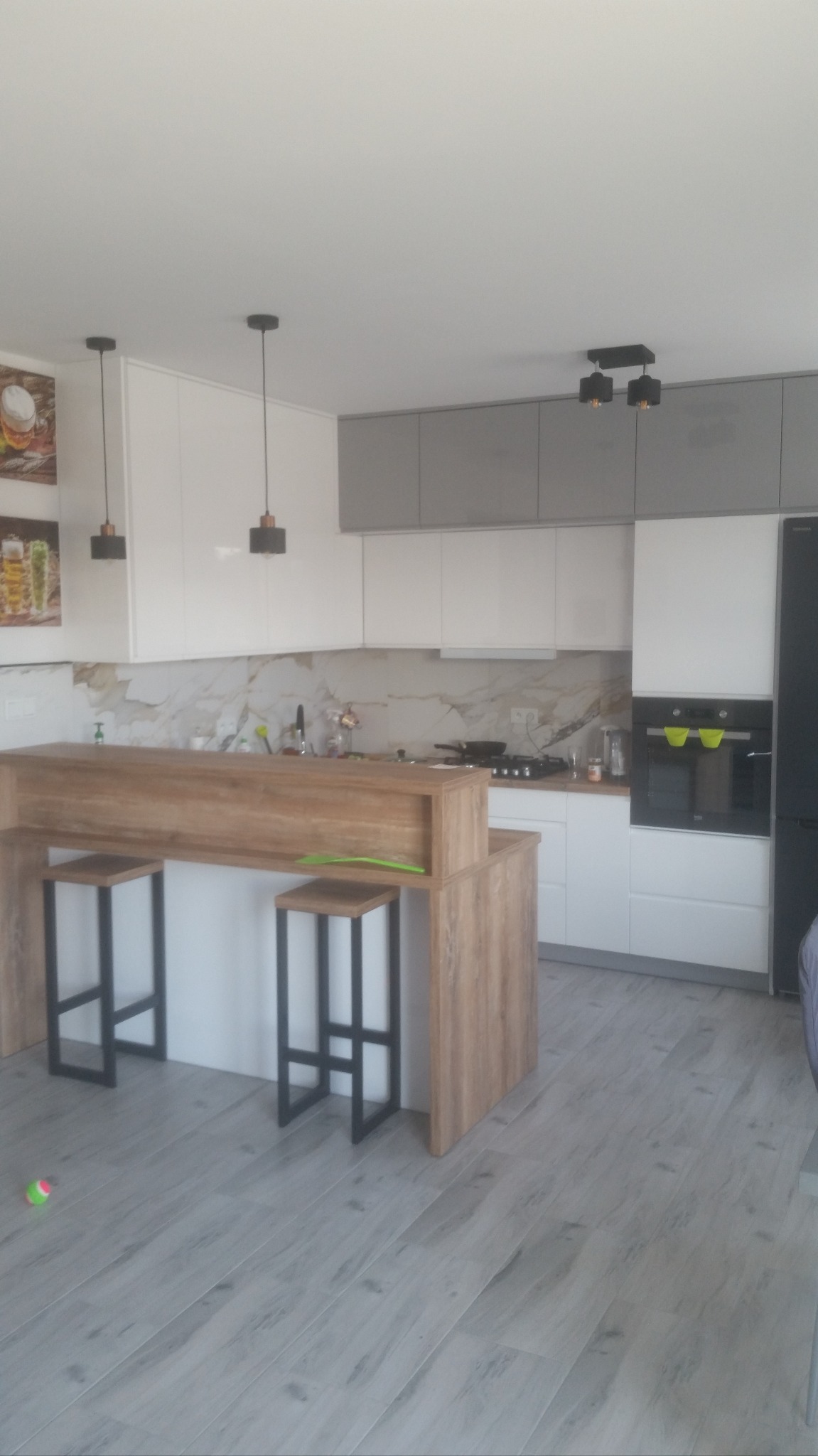 Nowoczesna kuchnia z wyspą i hokerami na metalowych nogach. Białe i szare szafki, marmurowy backsplash. Dwa wiszące światła nad wyspą. Jasna podłoga.