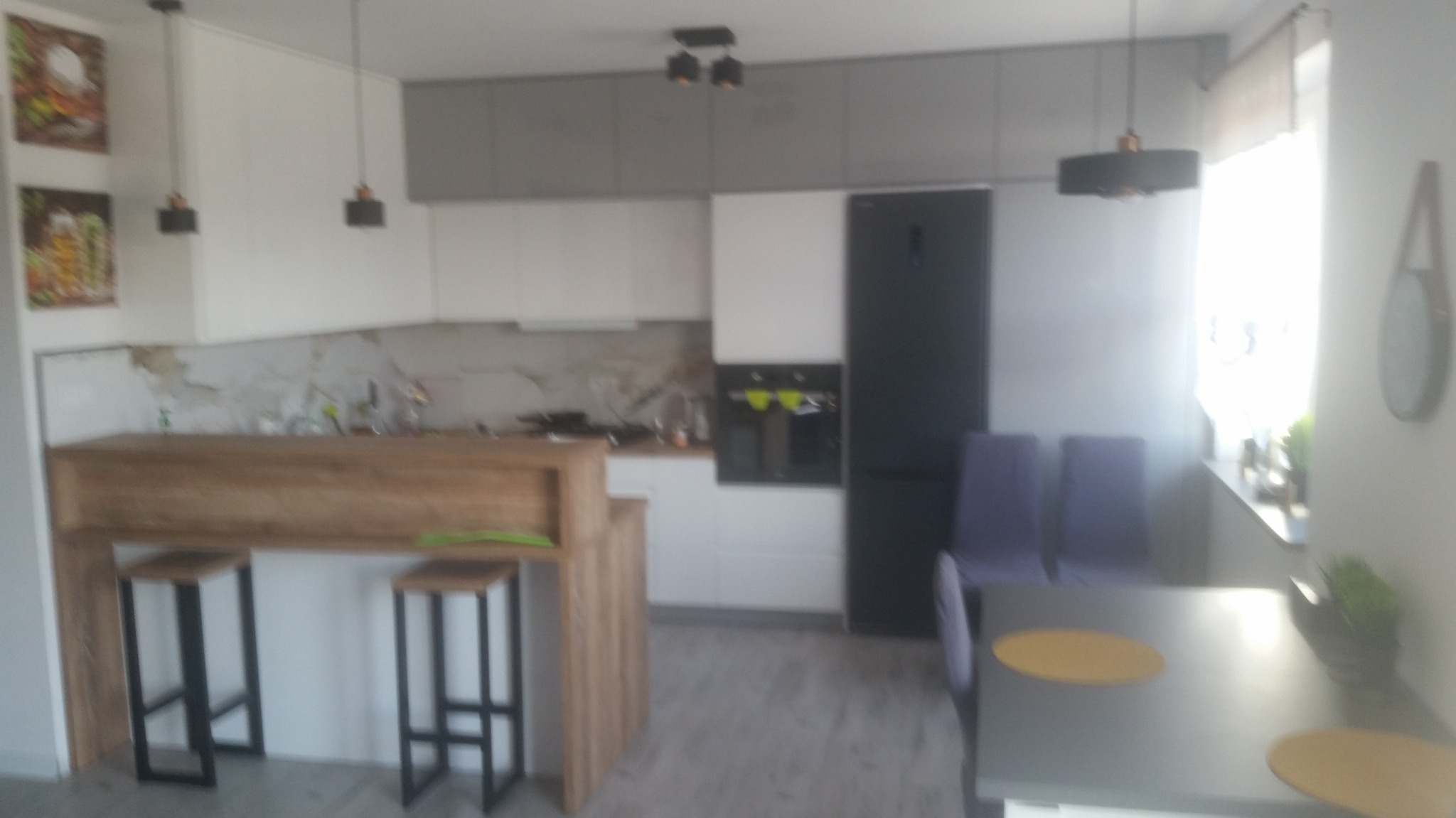 Nowoczesna kuchnia z drewnianym blatem barowym i hokerami. Białe i szare szafki, czarna lodówka. Dwa obrazy na ścianie. Minimalistyczne lampy wiszące.