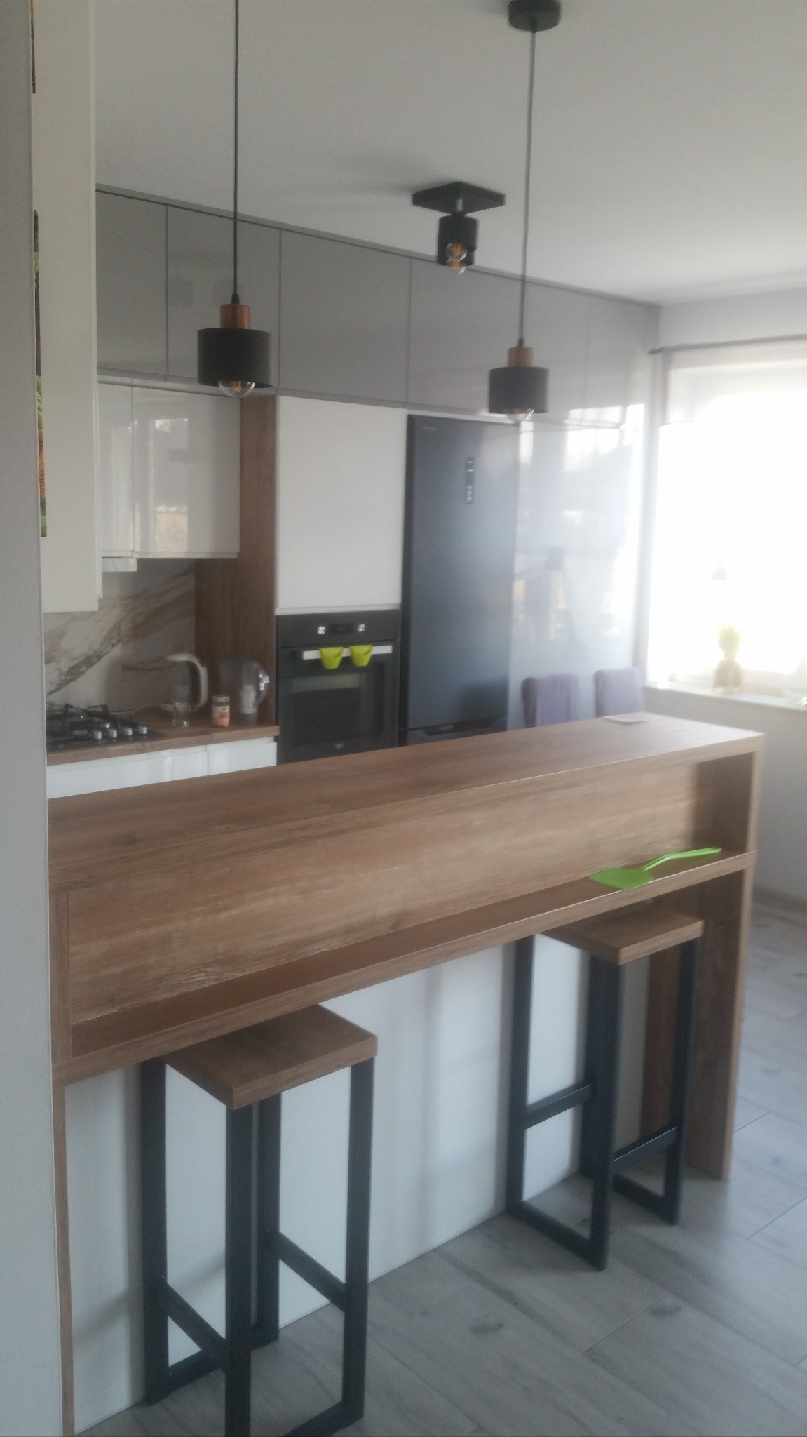 Nowoczesna kuchnia z drewnianym blatem barowym i hokerami na metalowych nogach. Szare szafki wiszące i białe dolne. Nad blatem wiszą czarne lampy.