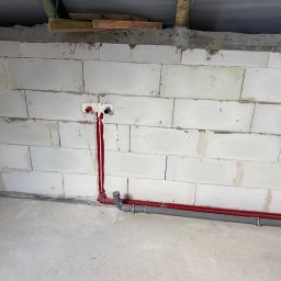 R.S.INSTAL Radosław Szczygieł - Świeżo położone, czerwone rury instalacji wodnej w surowym wnętrzu z białych bloczków, z widocznymi przyłączami i szarą rurą odpływową. Widoczne elementy konstrukcji dachu.