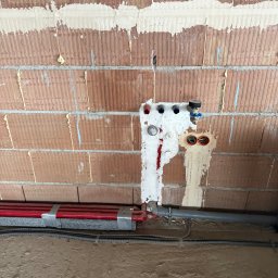 R.S.INSTAL Radosław Szczygieł - Czerwone rury grzewcze przy ścianie z cegły, zabezpieczone pianką montażową. Widoczne przyłącza i instalacje w trakcie montażu.