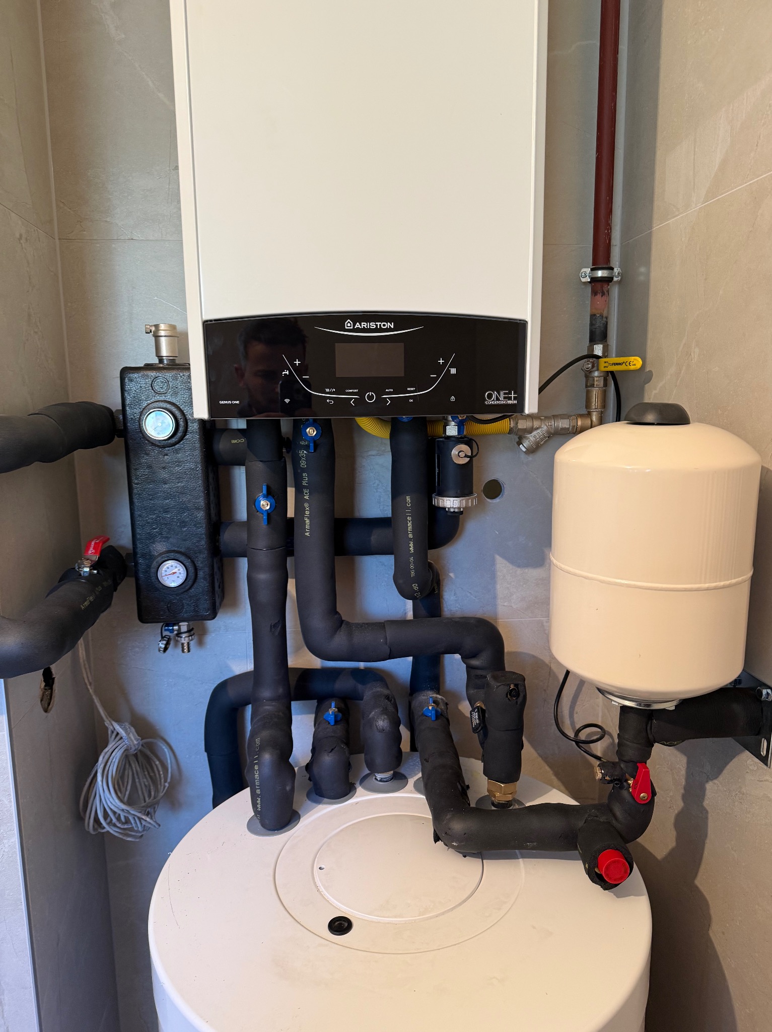 Nowoczesny, biały piec gazowy Ariston z czarnym panelem sterowania, podłączony do czarnych, izolowanych rur i białego zbiornika wyrównawczego na tle beżowych płytek.