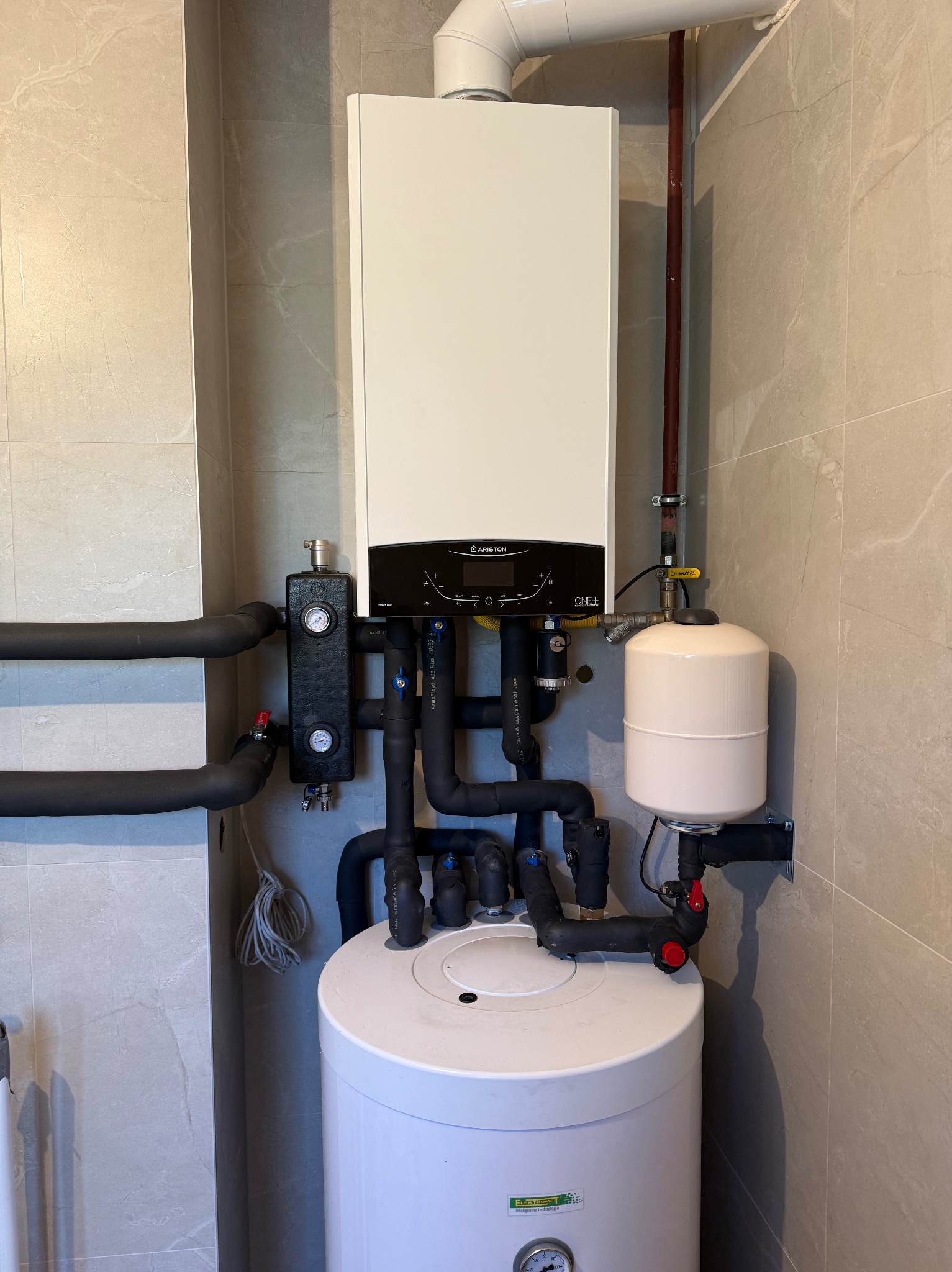 Nowoczesny, biały piec gazowy Ariston zamontowany na tle płytek imitujących marmur. Widoczne czarne rury, manometry i biały zbiornik. Instalacja w estetycznym wnętrzu.