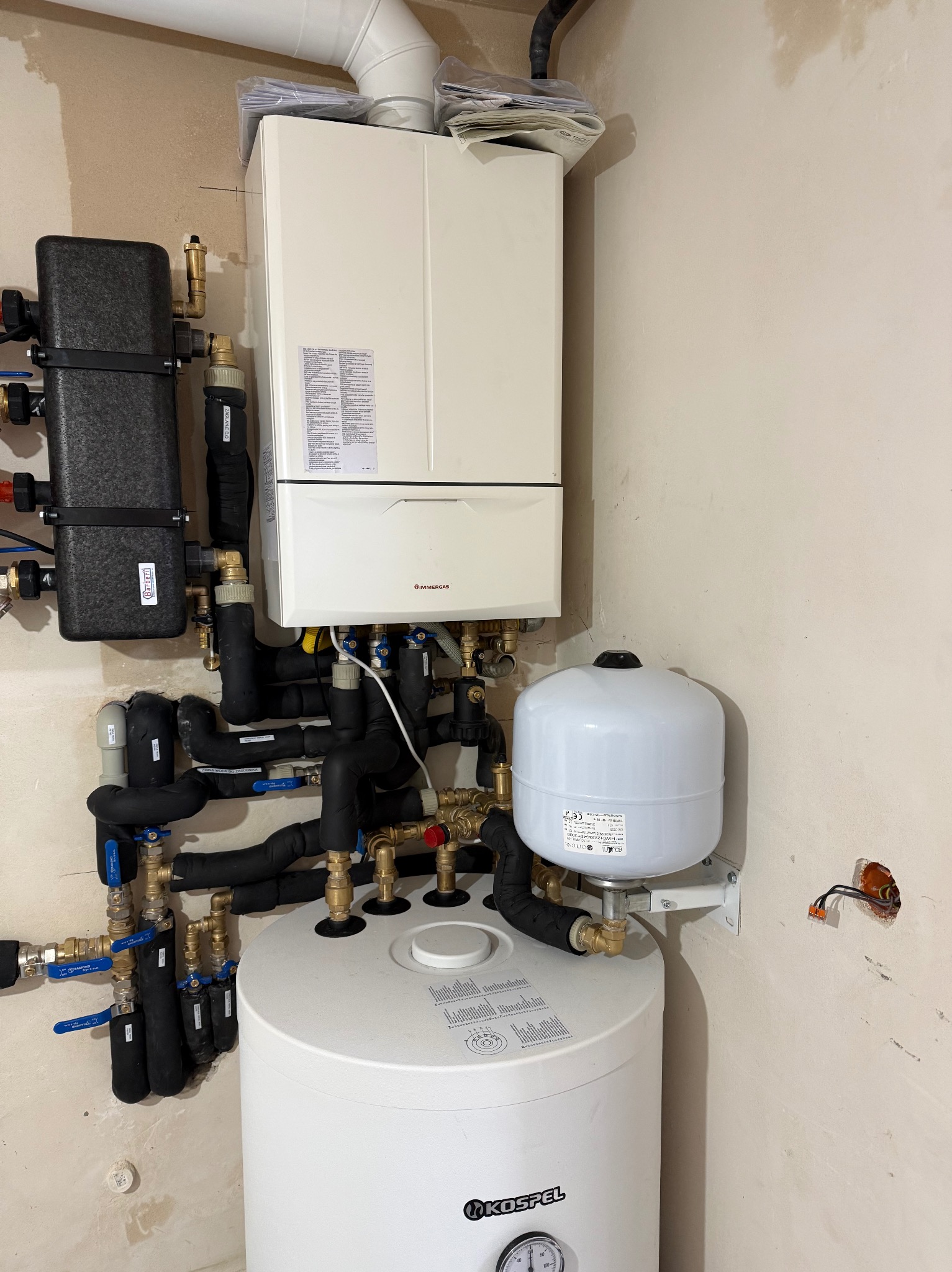 Kocioł gazowy Immergas z osprzętem, zbiornik Kospel, czarne rury z izolacją, zawory i kształtki mosiężne. Instalacja grzewcza w trakcie montażu.