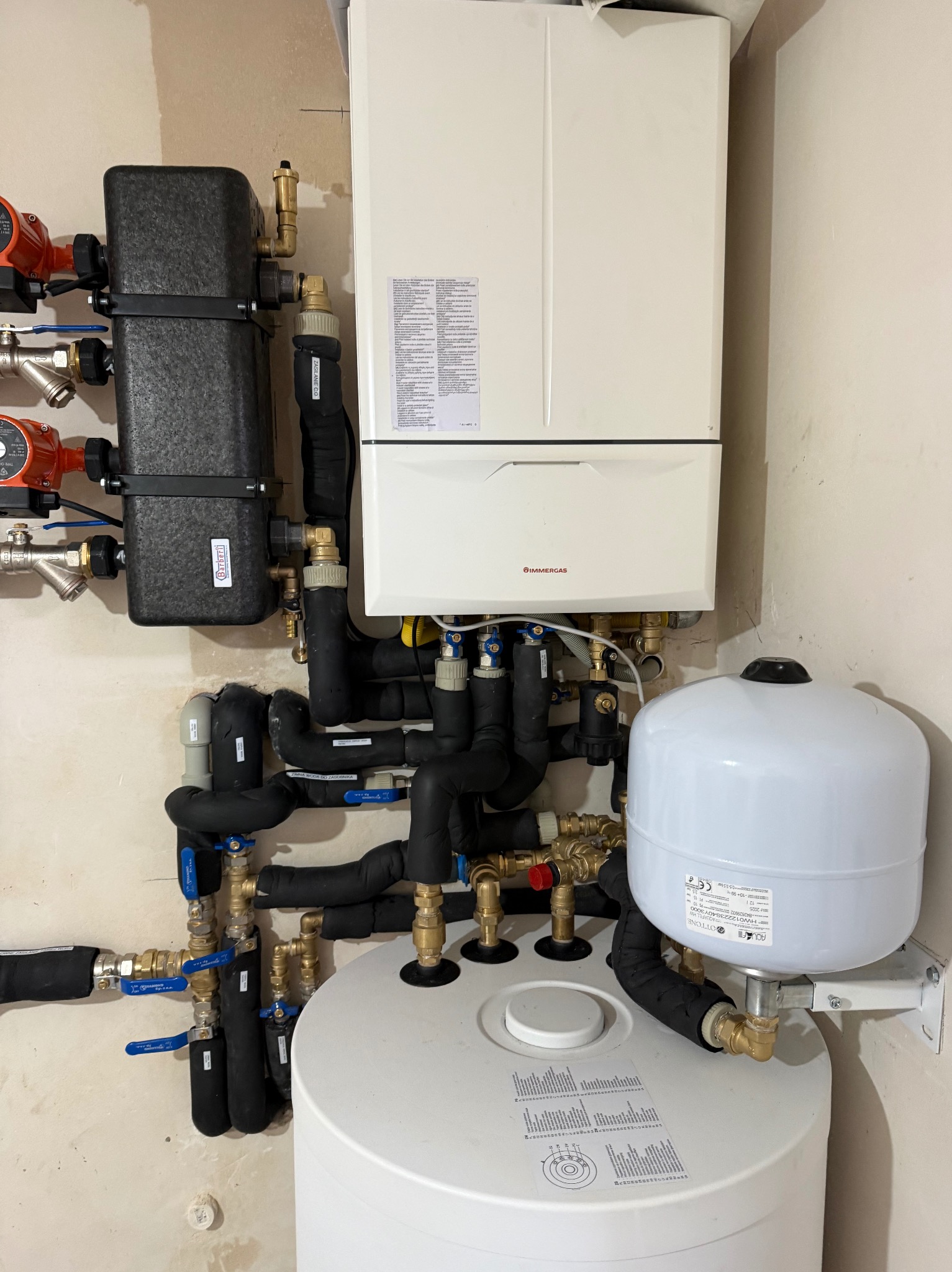 Nowoczesny piec gazowy Immergas z czarnymi rurami izolowanymi termicznie, zbiornikiem i pompami obiegowymi na tle jasnej ściany. Instalacja grzewcza w kotłowni.
