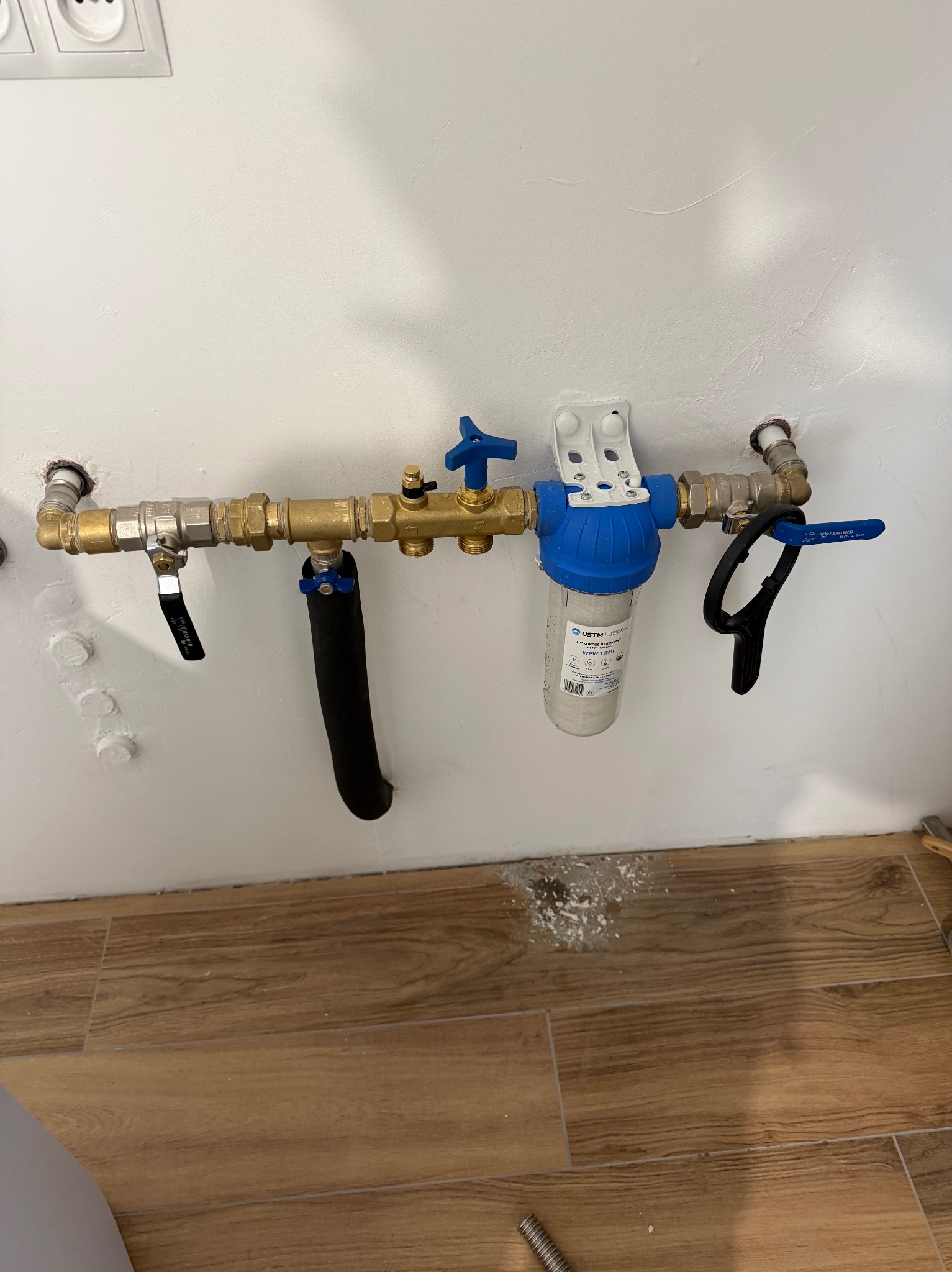 Zamontowany filtr wody USTM z niebieską obudową, połączony z mosiężnymi rurami i zaworami na białej ścianie, widoczne ślady po wierceniu na podłodze.