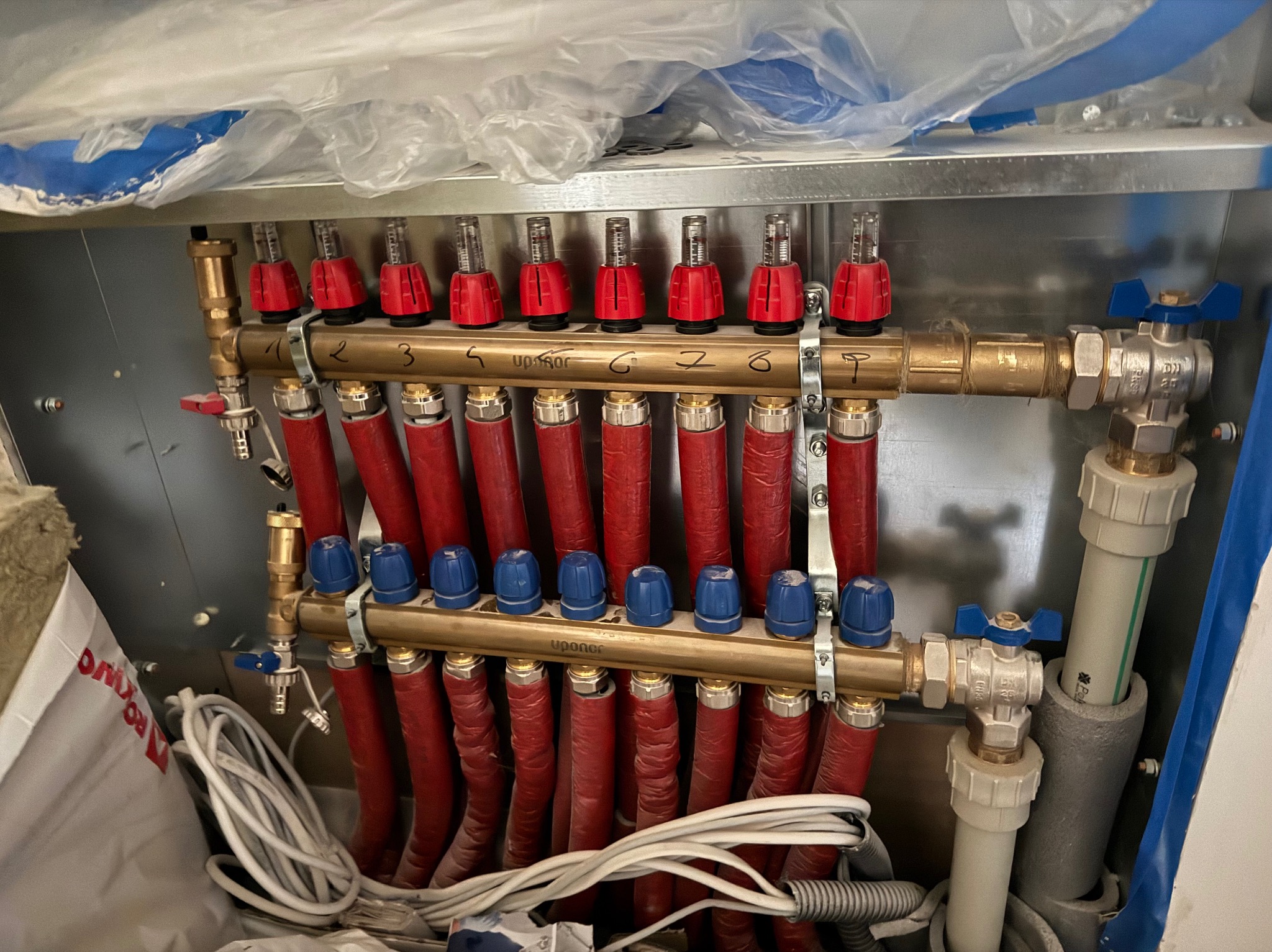 Rozdzielacz ogrzewania podłogowego z czerwonymi rurami i zaworami z niebieskimi pokrętłami, zamontowany w metalowej szafce, widoczne przewody elektryczne i izolacja.