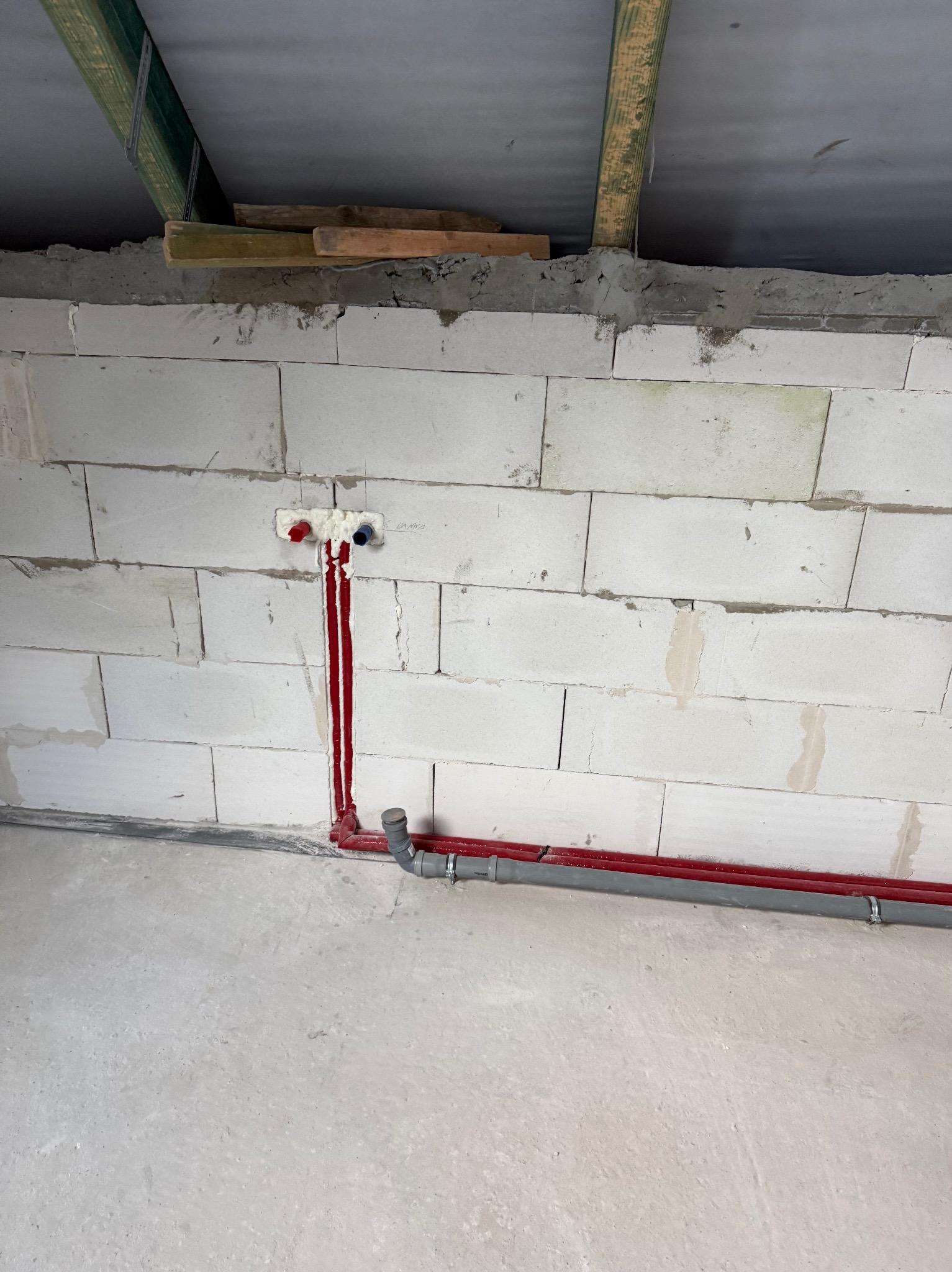 Świeżo położone, czerwone rury instalacji wodnej w surowym wnętrzu z białych bloczków, z widocznymi przyłączami i szarą rurą odpływową. Widoczne elementy konstrukcji dachu.
