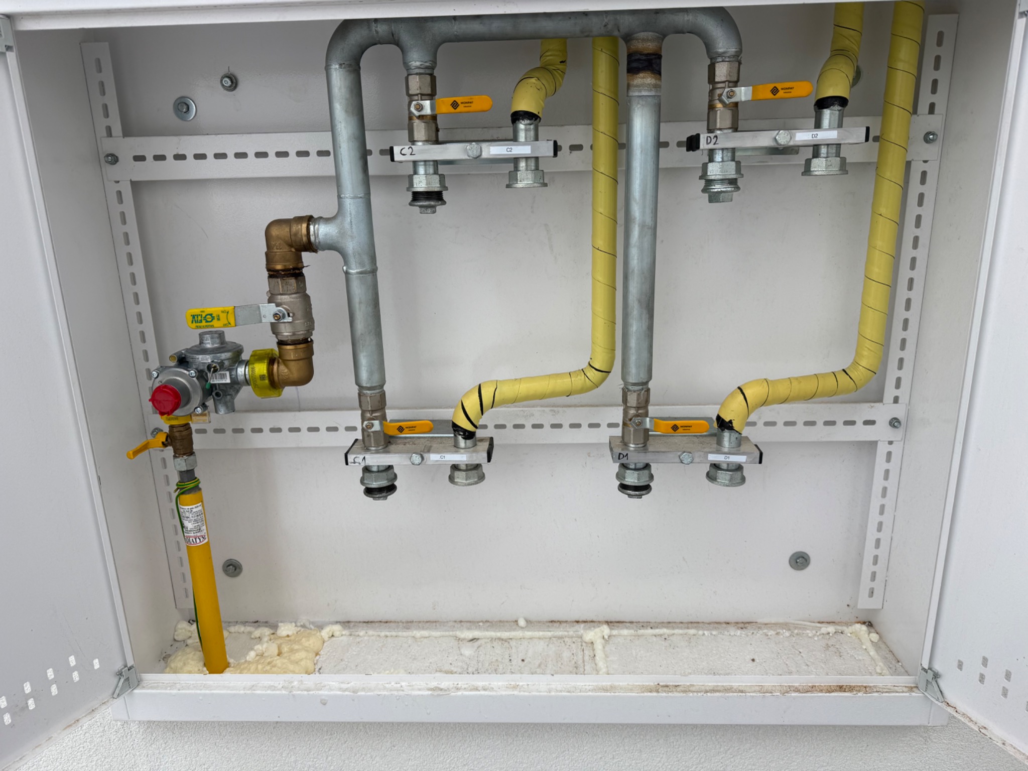 Węzeł gazowy w białej szafce: żółte rury w izolacji, zawory z żółtymi uchwytami, metalowe rury i mocowania. Widoczne oznaczenia C1, C2, D1, D2. Pianka montażowa u dołu.
