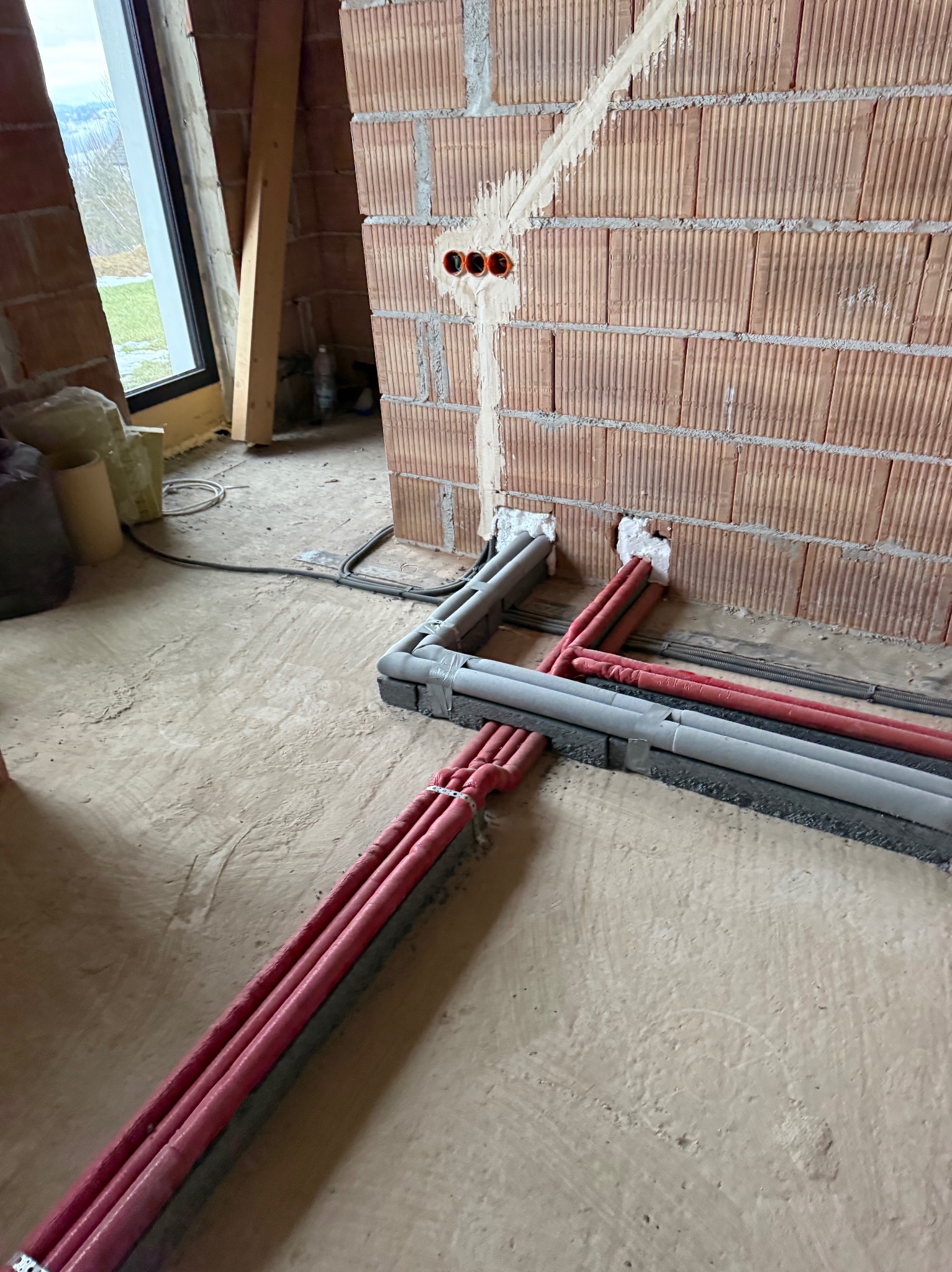 Instalacja grzewcza w trakcie montażu: czerwone rury w szarych osłonach prowadzone wzdłuż ściany z cegły dziurawki. Widoczne puszki elektryczne i okno.