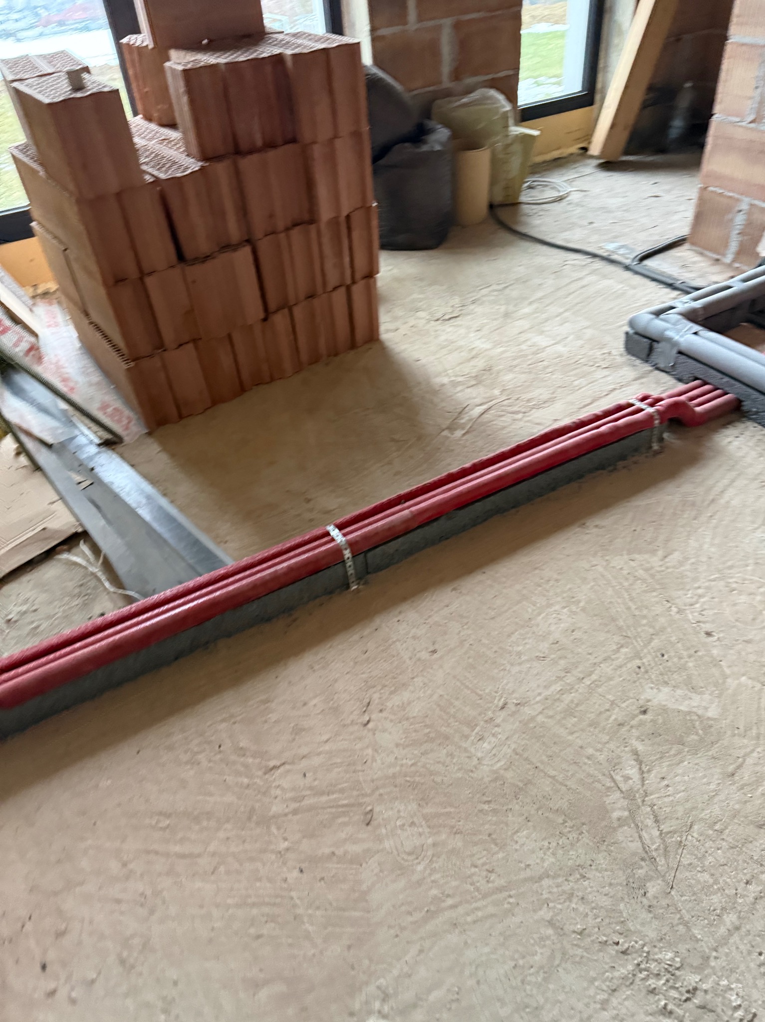 Czerwone rury w szarej osłonie, zabezpieczone metalową obejmą, leżą na surowej posadzce w trakcie budowy. W tle cegły i okno.