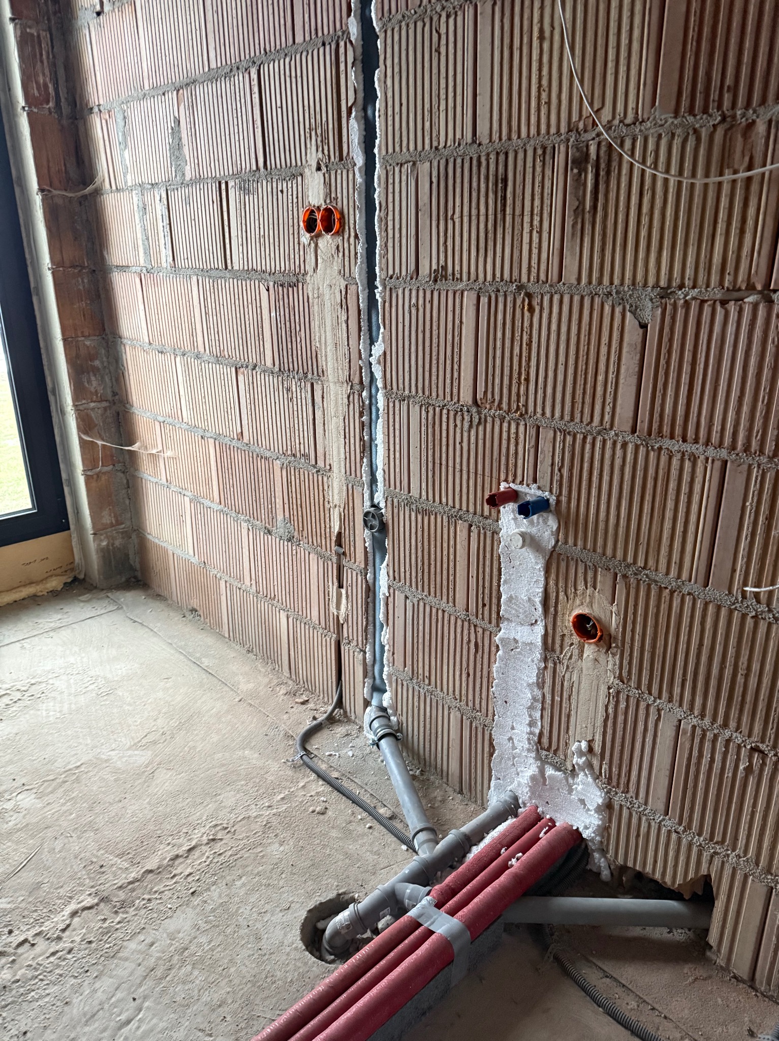 Instalacja wodna w bruzdach ściany z czerwonej cegły, zabezpieczona pianką montażową. Widoczne rury szare i czerwone. Przygotowanie do dalszych prac wykończeniowych.