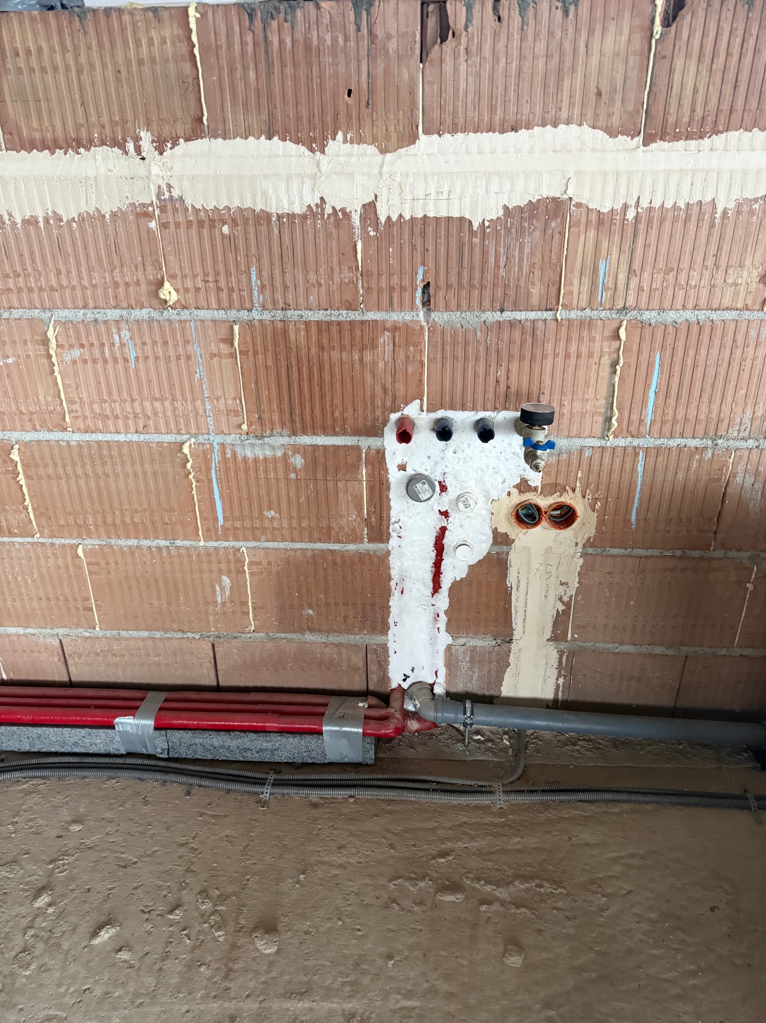Czerwone rury grzewcze przy ścianie z cegły, zabezpieczone pianką montażową. Widoczne przyłącza i instalacje w trakcie montażu.