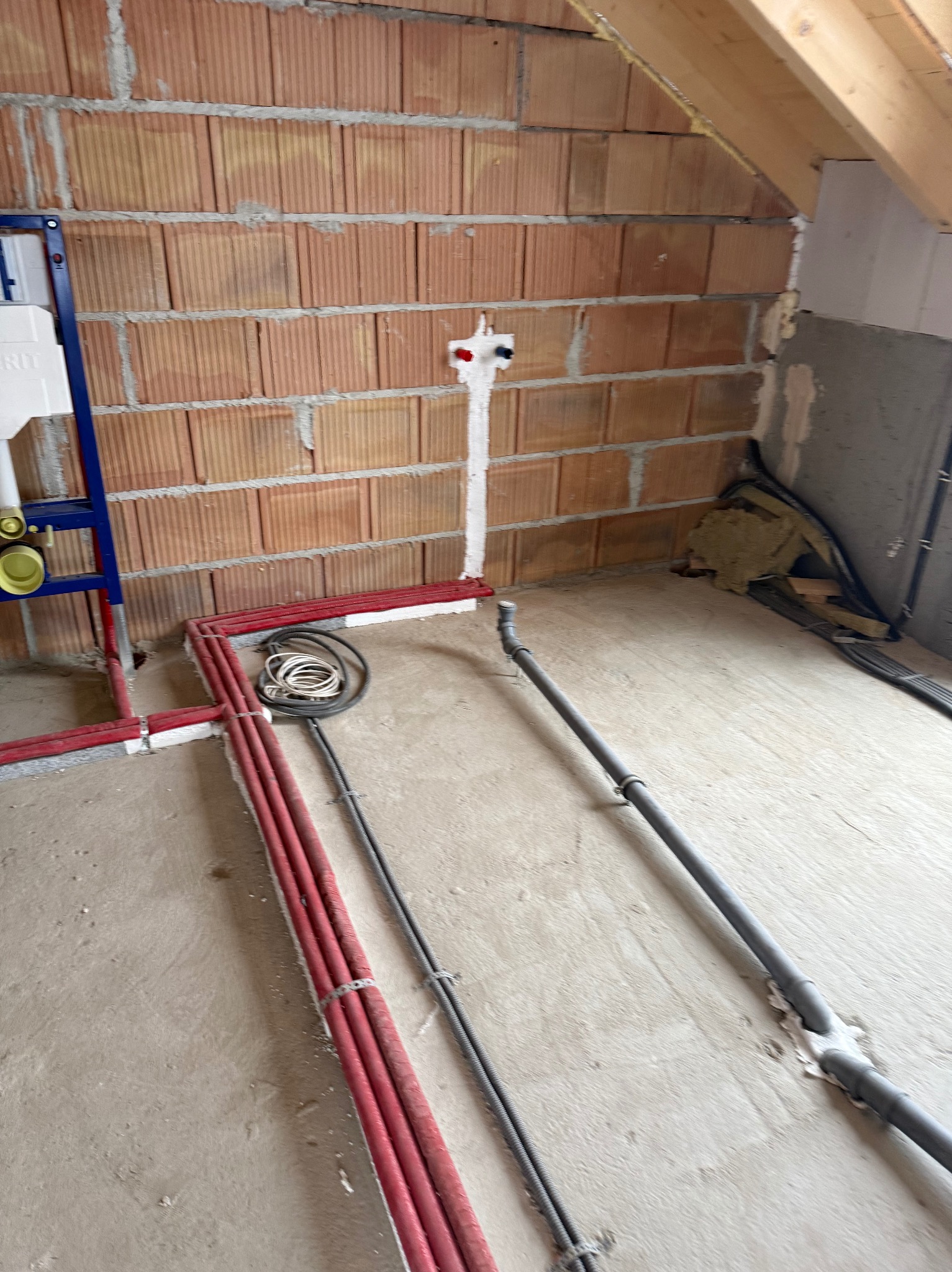 Czerwone rury instalacji CO ułożone w surowym wnętrzu z ceglaną ścianą i elementami poddasza. Widoczne także szare rury kanalizacyjne i niebieski stelaż podtynkowy WC.