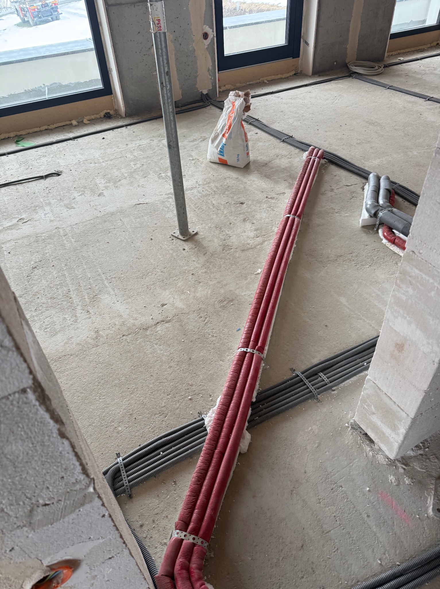 Wnętrze w trakcie budowy z ułożonymi rurami grzewczymi w izolacji i szarymi rurami karbowanymi, widoczne okna i worek z zaprawą.