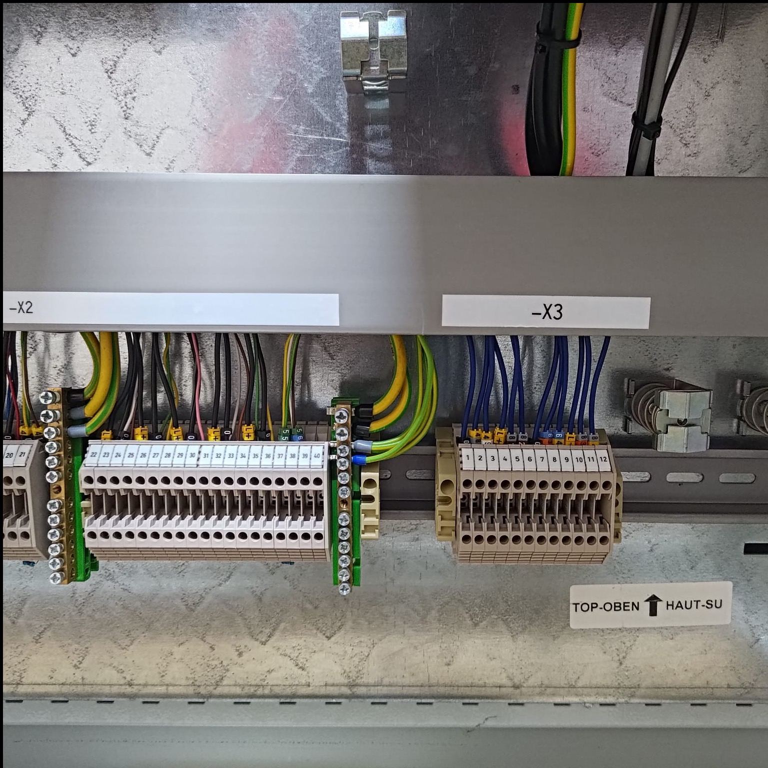 Rozdzielnica elektryczna z podłączonymi przewodami o różnych kolorach, oznaczona etykietami -X2 i -X3. Widoczne złącza i listwy zaciskowe. Metalowa obudowa.