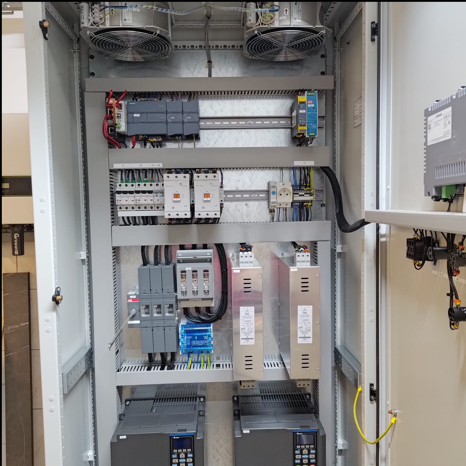 Wnętrze szafy sterowniczej z licznymi elementami elektrycznymi, w tym wentylatory, sterowniki PLC, zasilacze i zabezpieczenia. Widoczne okablowanie i uziemienie.