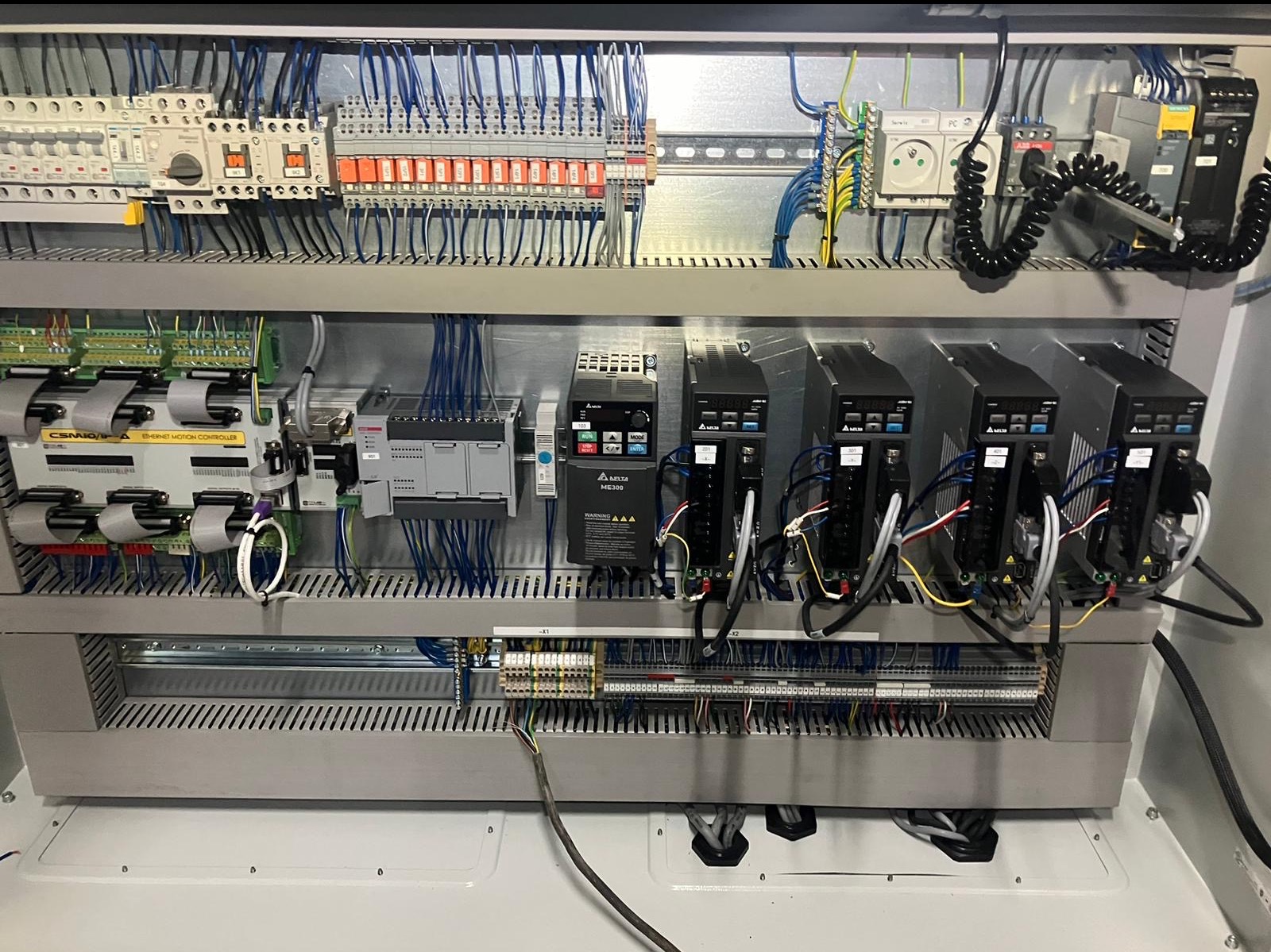 Rozbudowana szafa sterownicza z licznymi modułami, przewodami i złączami. Widoczne sterowniki silników Delta ME300 i inne komponenty. Precyzyjne okablowanie wewnątrz metalowej obudowy.
