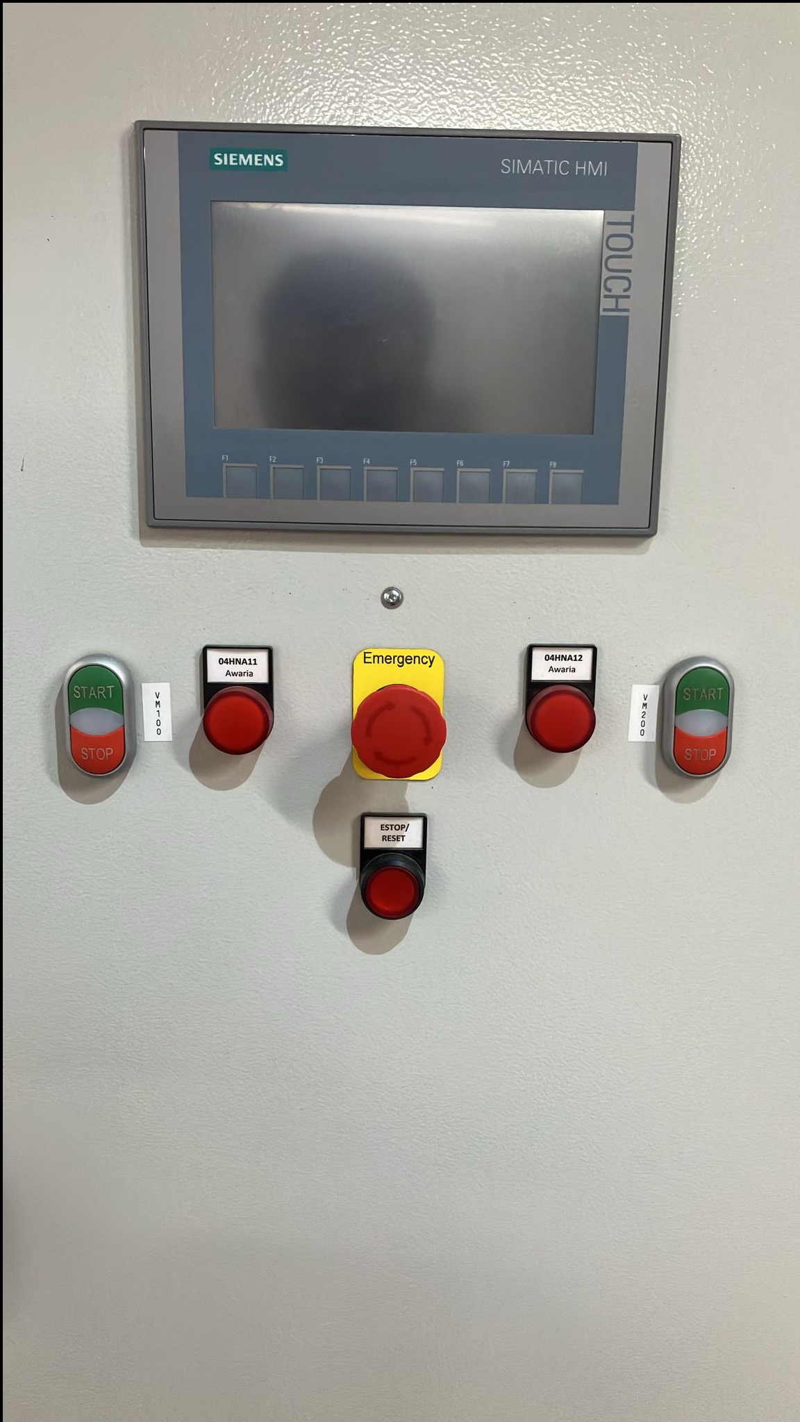 Panel sterowania Siemens Simatic HMI z przyciskami awaryjnymi, start/stop i reset. Widoczne oznaczenia i etykiety. Szary panel na teksturowanej powierzchni.