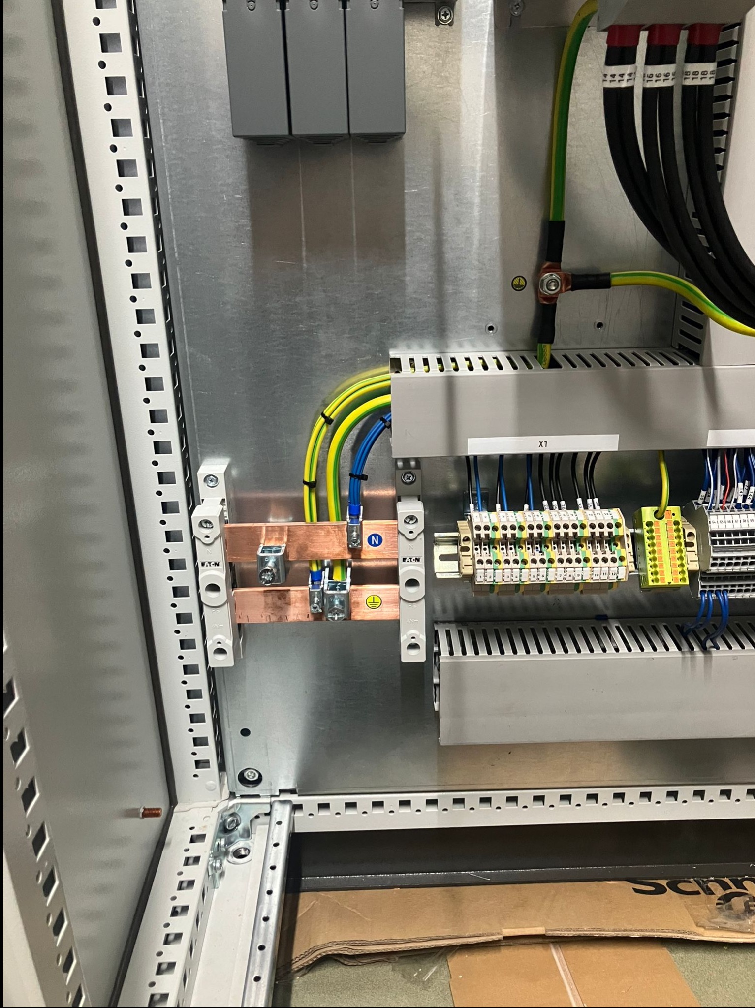 Wnętrze szafy sterowniczej z widocznymi elementami instalacji elektrycznej: szyny, przewody w kolorach żółto-zielonym i niebieskim, złączki. Widoczne oznaczenia na przewodach.