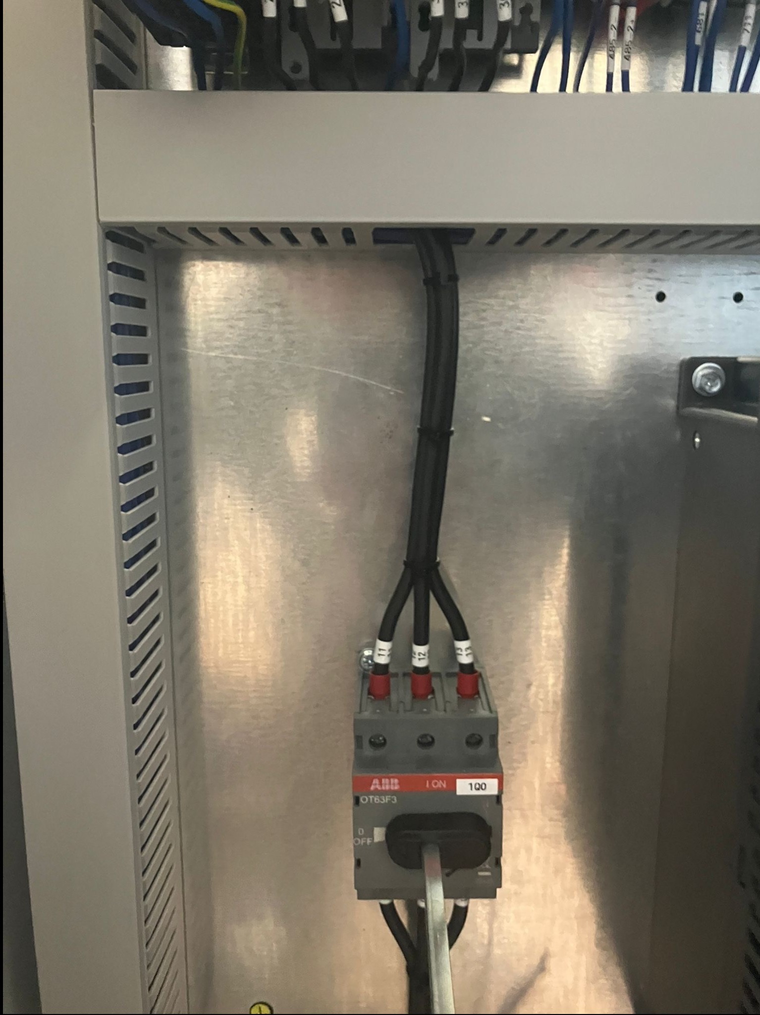 Szczegółowe ujęcie wnętrza szafy sterowniczej z widocznym wyłącznikiem ABB OT63F3 i podłączonymi przewodami. Metalowa obudowa z perforacją po lewej stronie.