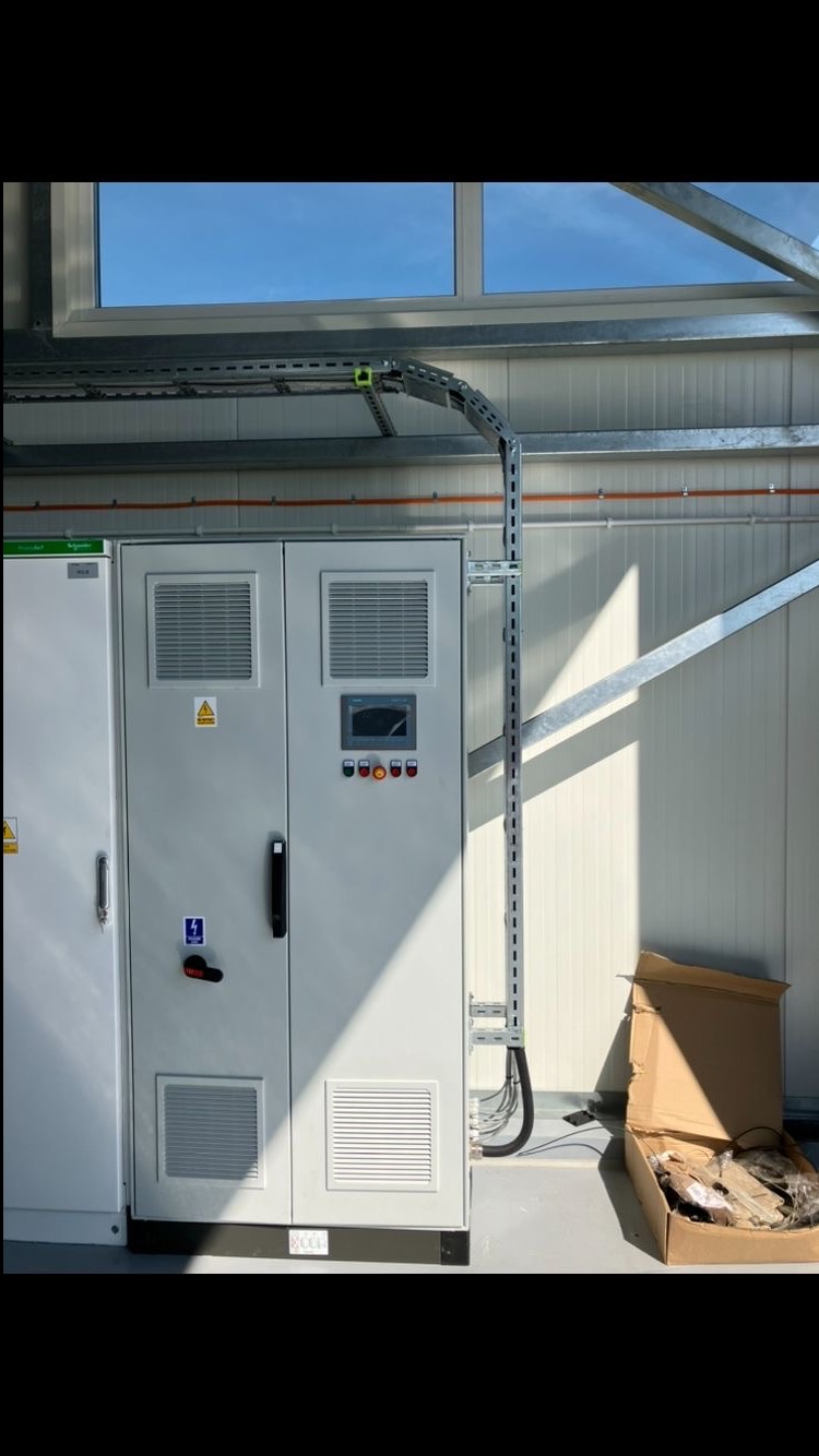 Szara szafa elektryczna z kratkami wentylacyjnymi i oznaczeniami ostrzegawczymi, z metalową drabinką kablową i przewodami w tle, w hali z oknem i kartonem.