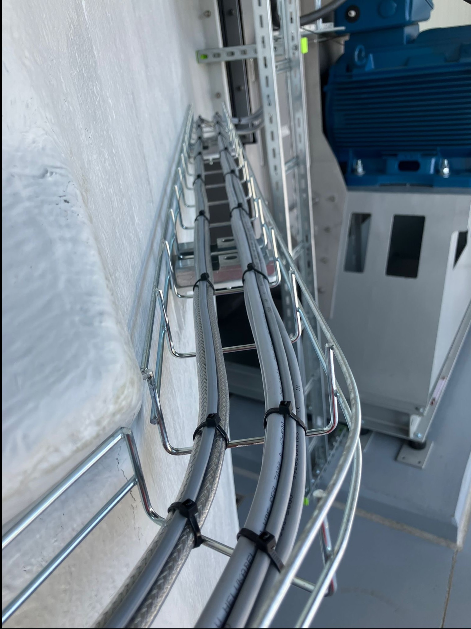 Szara metalowa korytka kablowa z przymocowanymi przewodami, zamocowana na białej ścianie. W tle widoczny niebieski silnik elektryczny.