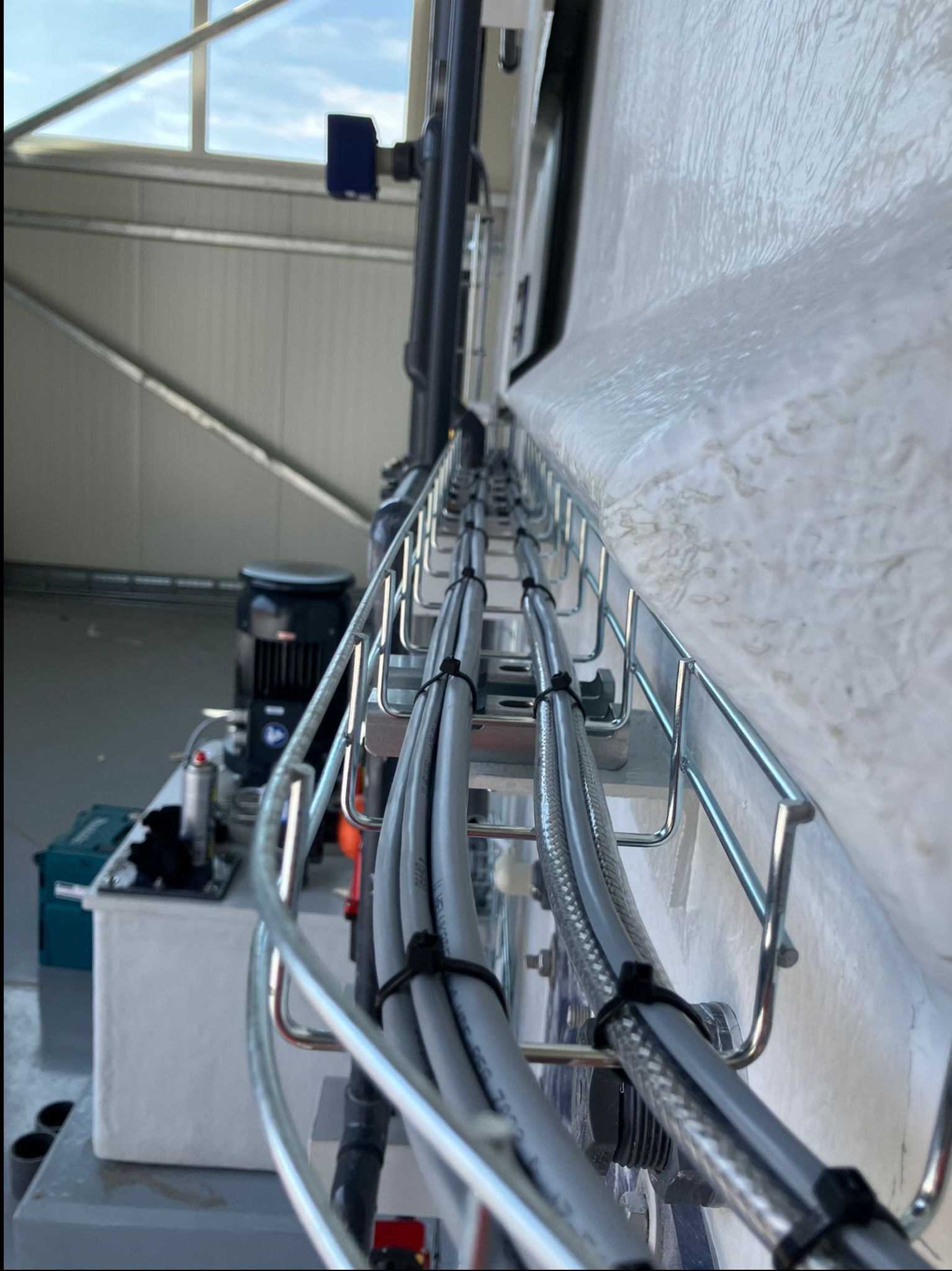 Szara instalacja kablowa w metalowej korycie, przymocowana do białej ściany w hali przemysłowej. Widoczne przewody i elementy mocujące.