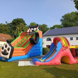 AMW Zjeżdżalnie Dmuchane - Dwa kolorowe dmuchańce na trawie: jeden z motywem kreskówkowym, drugi zjeżdżalnia z tunelem. W tle budynek z szarą dachówką i drzewa.